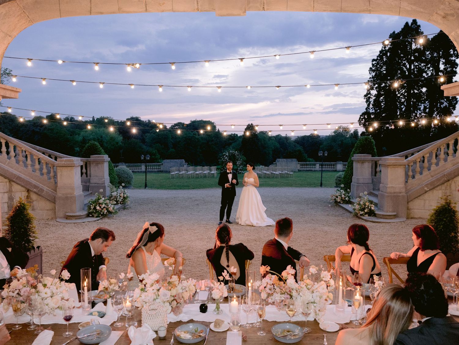 Elegant outdoor wedding reception under string lights with long tables set for dining at sunset.
