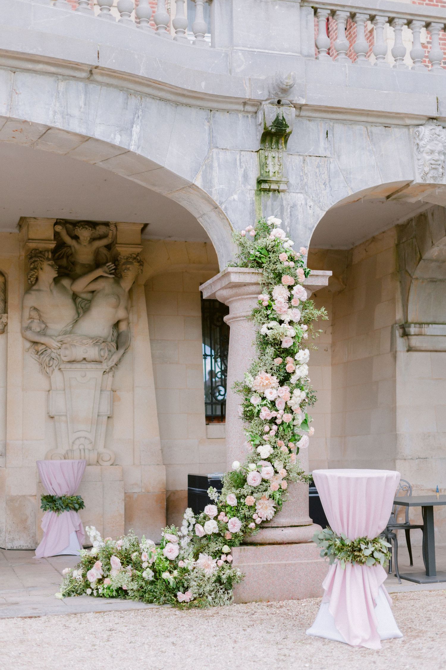Outdoor wedding venue with white floral arrangements and pink cocktail tables under stone archways.