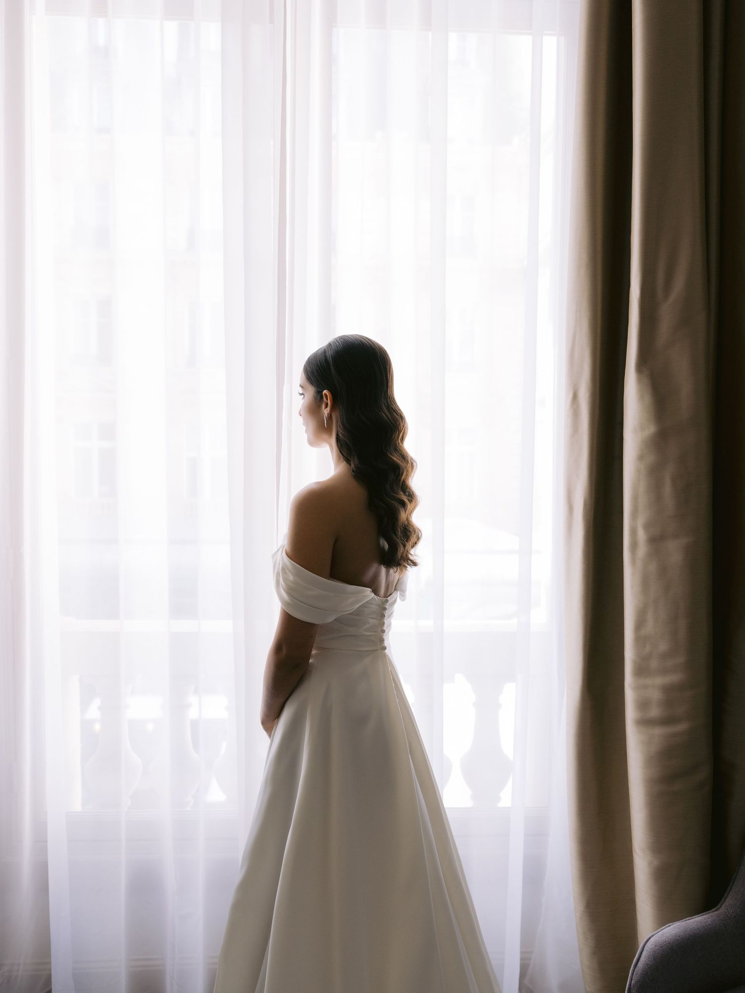 A bride in a white dress stands silhouetted against bright window light.