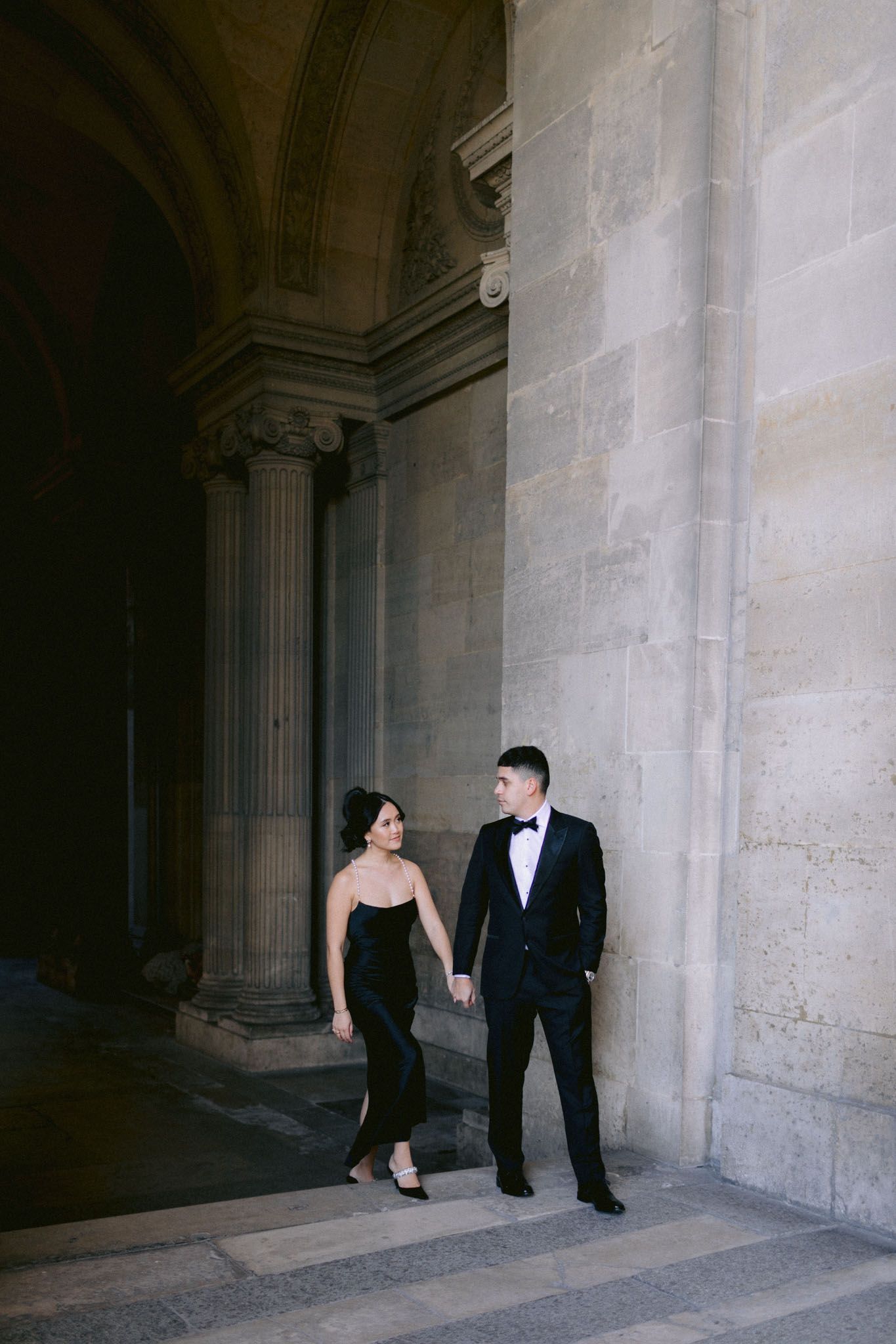 Couple in formal attire posing against classical stone architecture with columns.