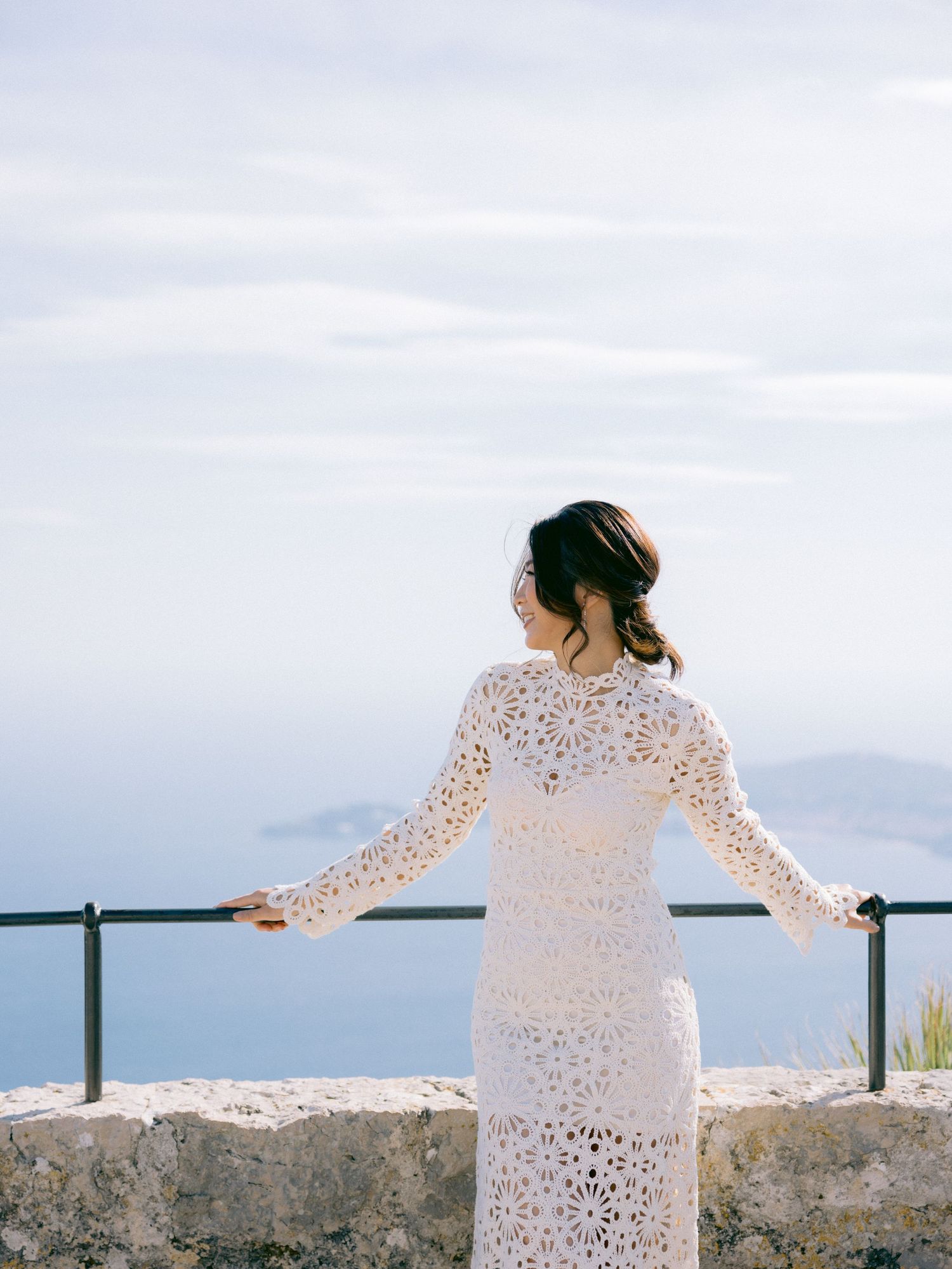 A bride in a delicate lace dress gazes at the scenic mountain view from a stone overlook.