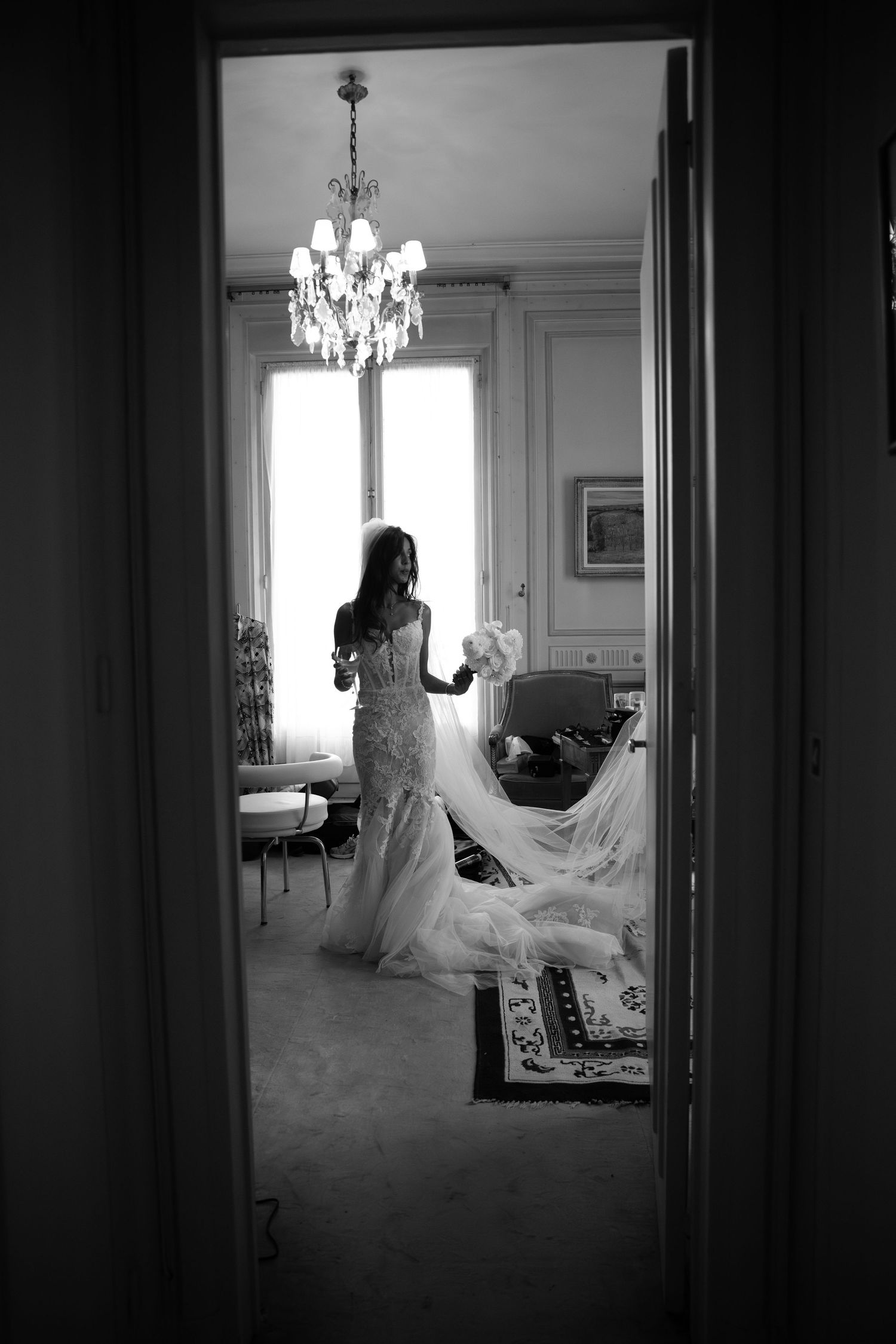 A silhouette through a doorway shows a bride in her gown beneath a crystal chandelier.