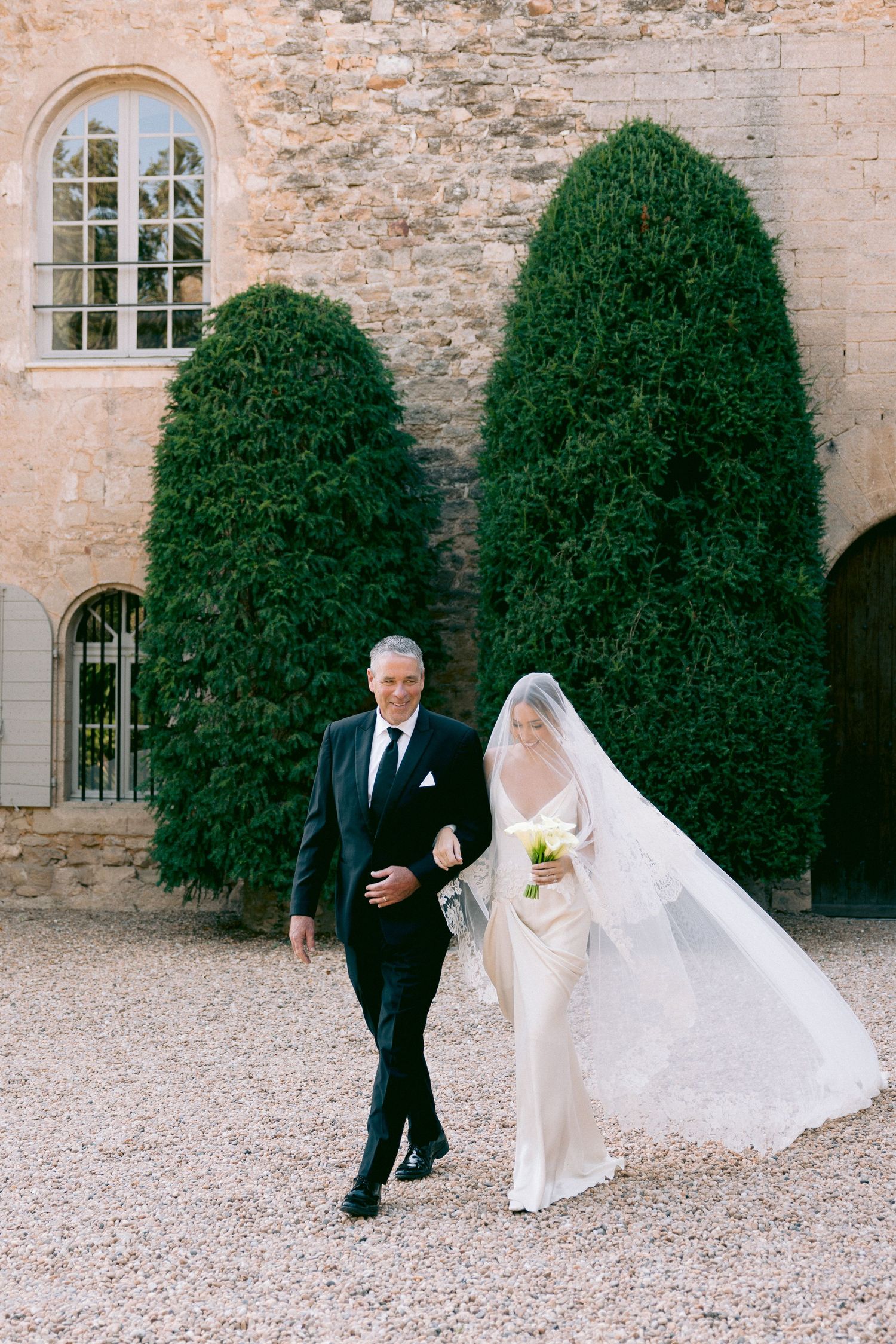 Elegant wedding ceremony setup against stone wall with manicured topiary bushes and arched windows.