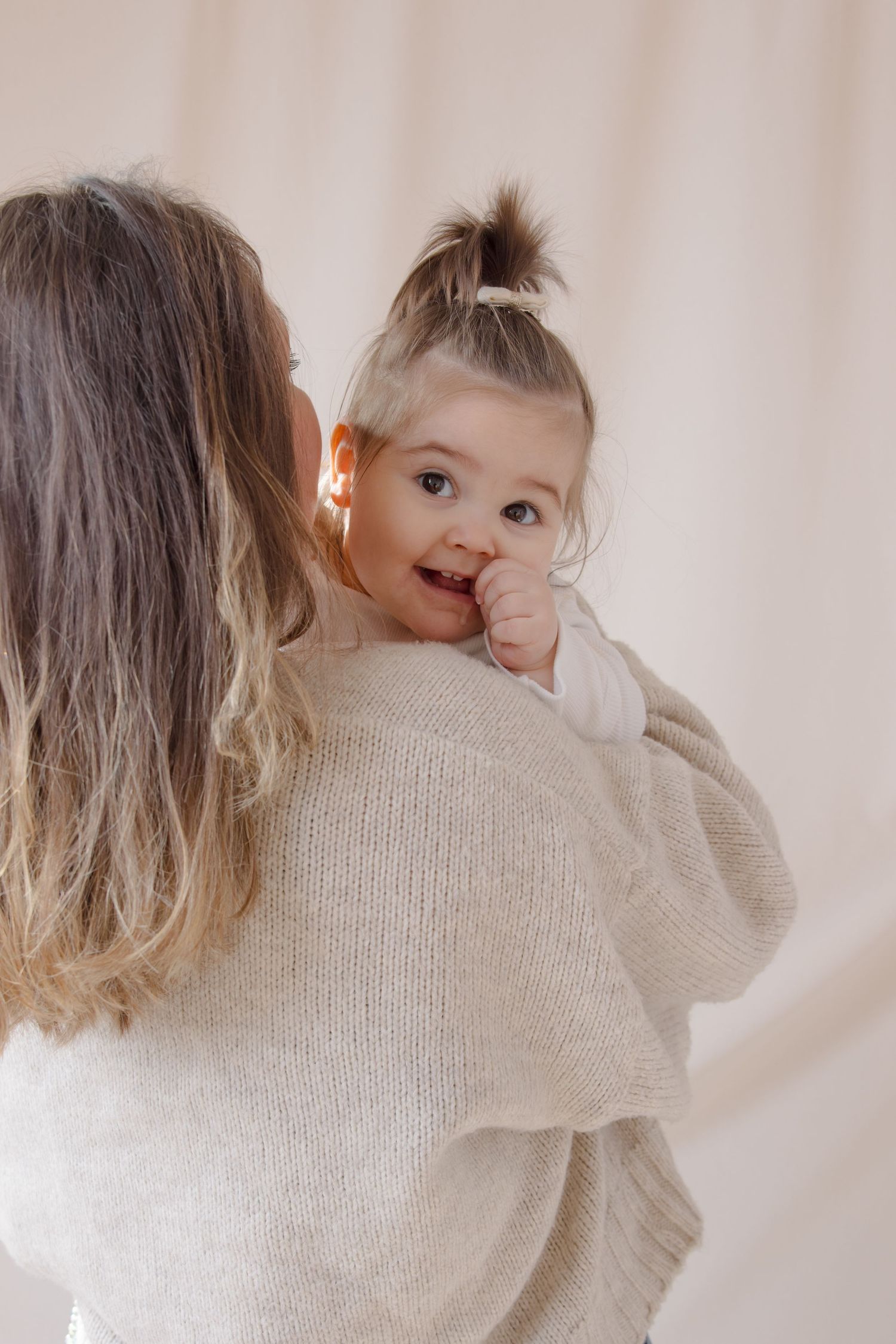 A mother holding her smiling baby girl with a tiny ponytail, both wearing cozy cream sweaters.