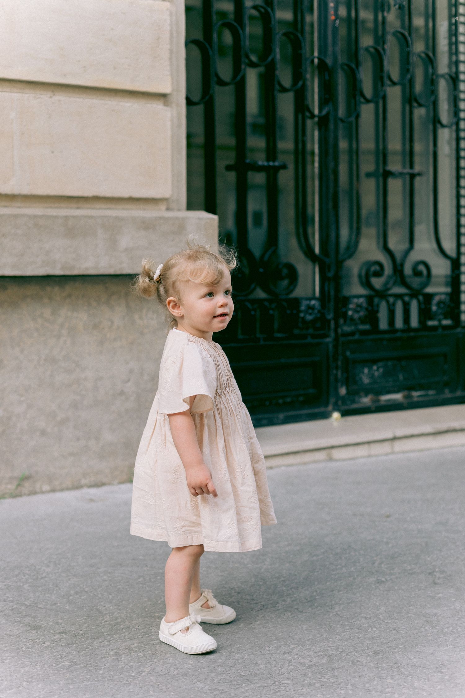 A small child in a pink dress walking along a stone building with iron gates.