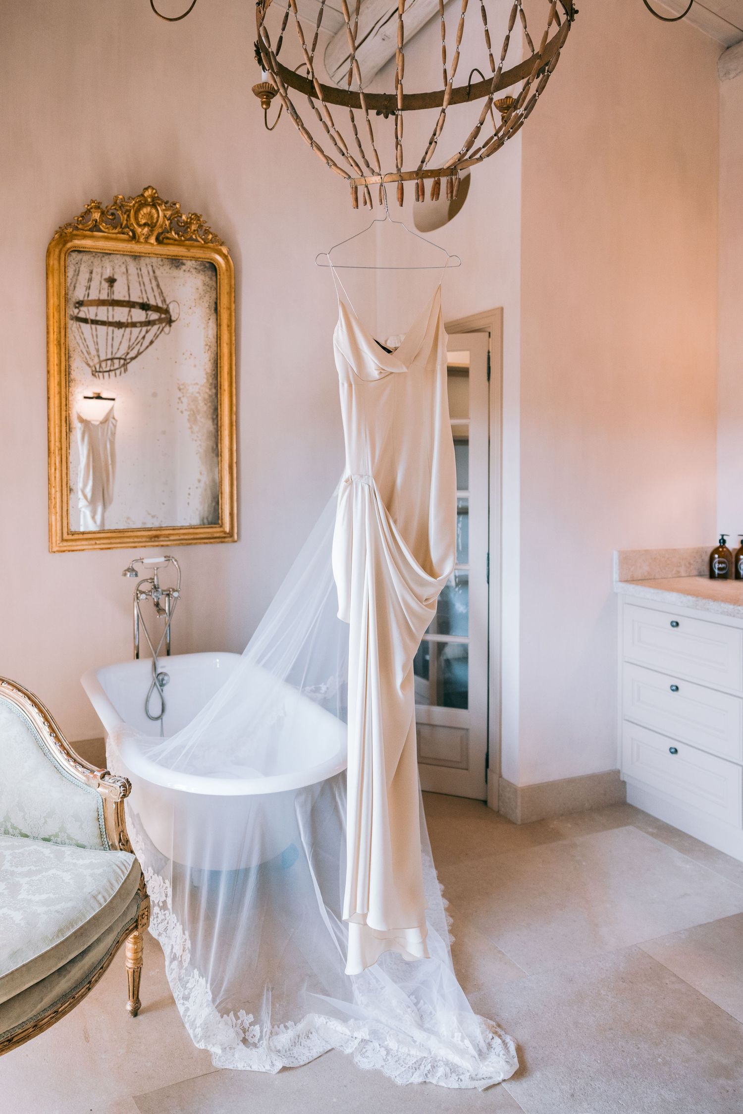 Ethereal wedding dress displayed in an elegant room with ornate gold mirror and chandelier.