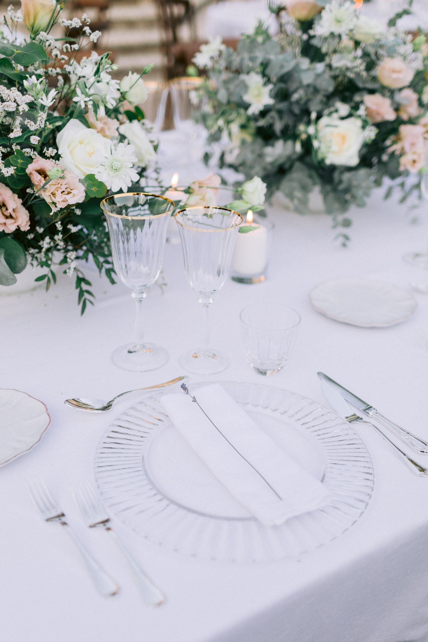 Close-up of elegant place settings with fluted glass plates and floral arrangements on white tablecloths.