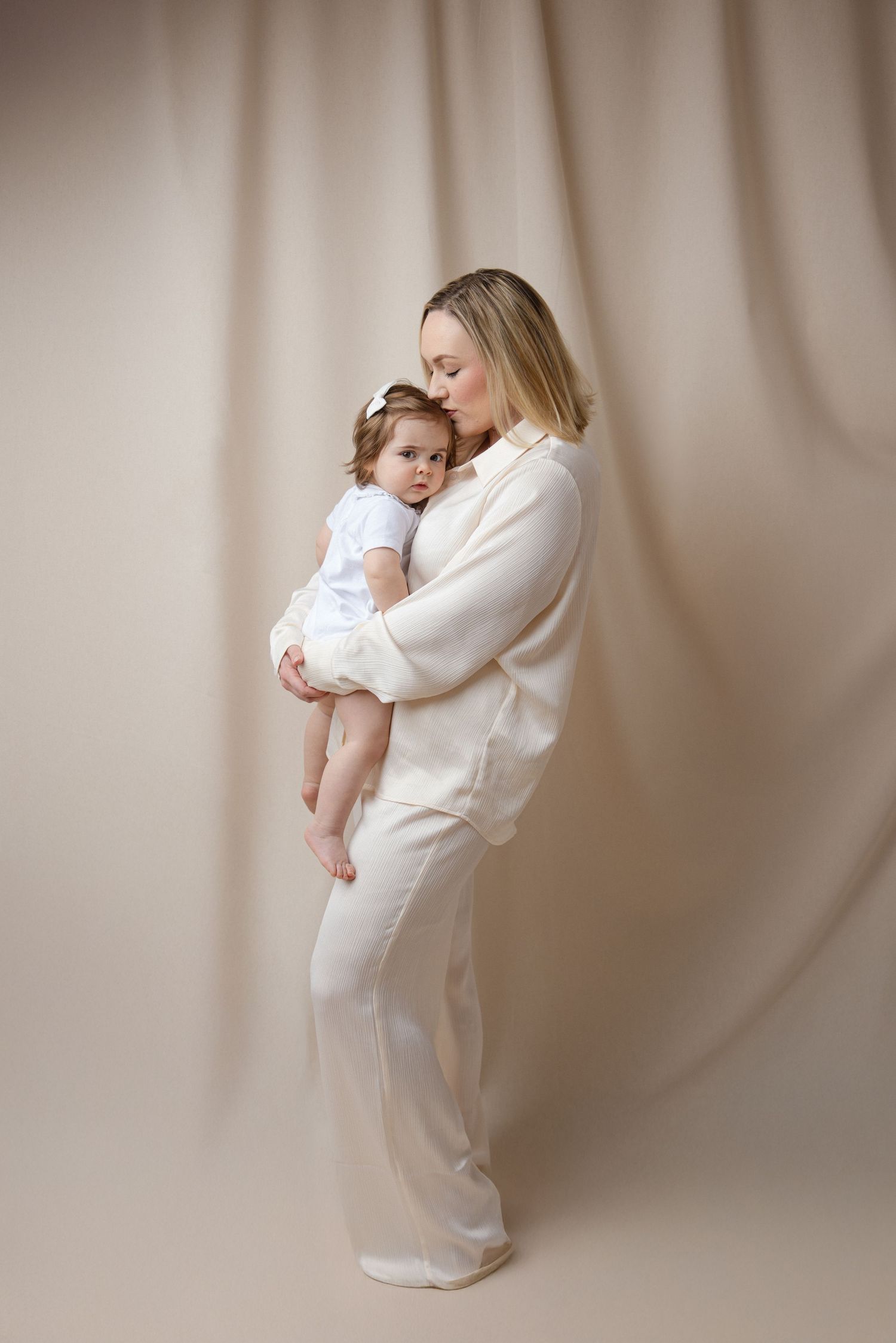 A mother in a cream outfit tenderly holds her baby girl in a white dress against a soft beige backdrop.
