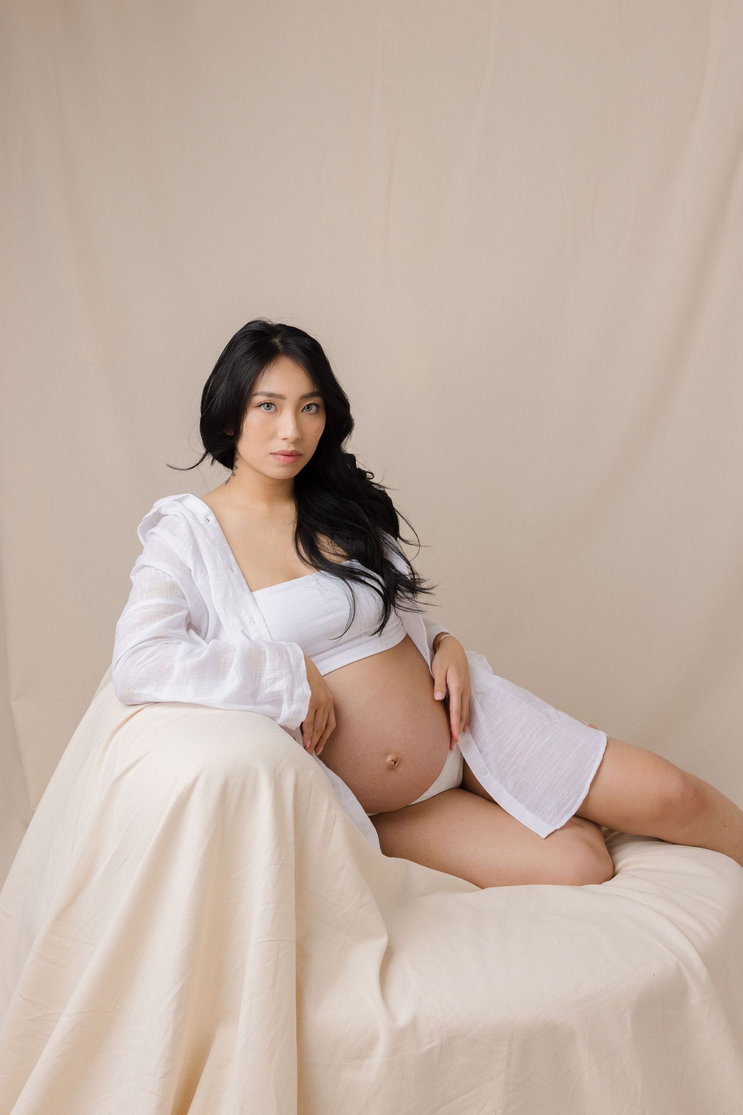Pregnant woman in white lingerie and open shirt poses gracefully on draped furniture against a neutral beige backdrop.