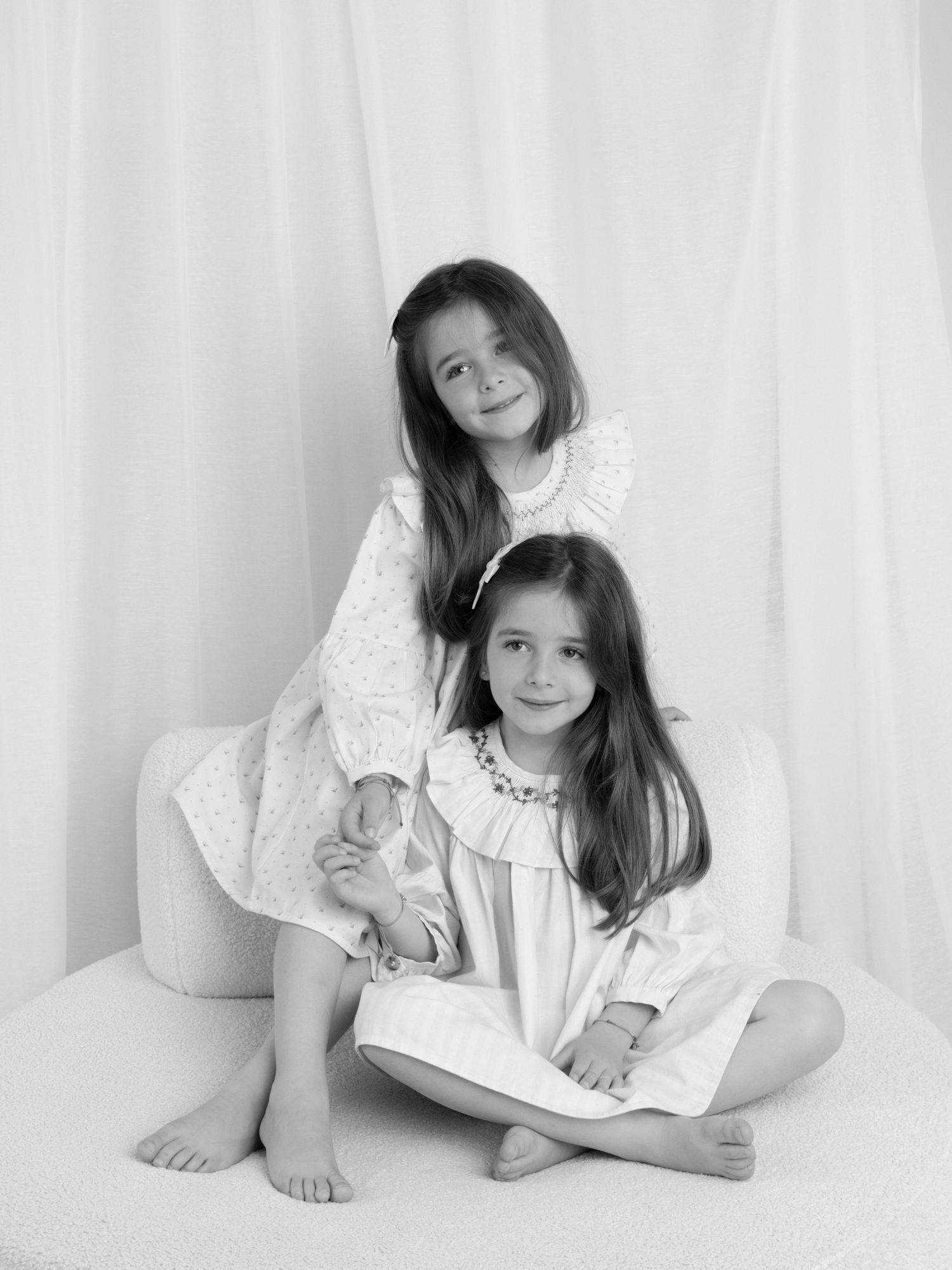 Black and white portrait of two young sisters in white dresses, posing together on a white chair.
