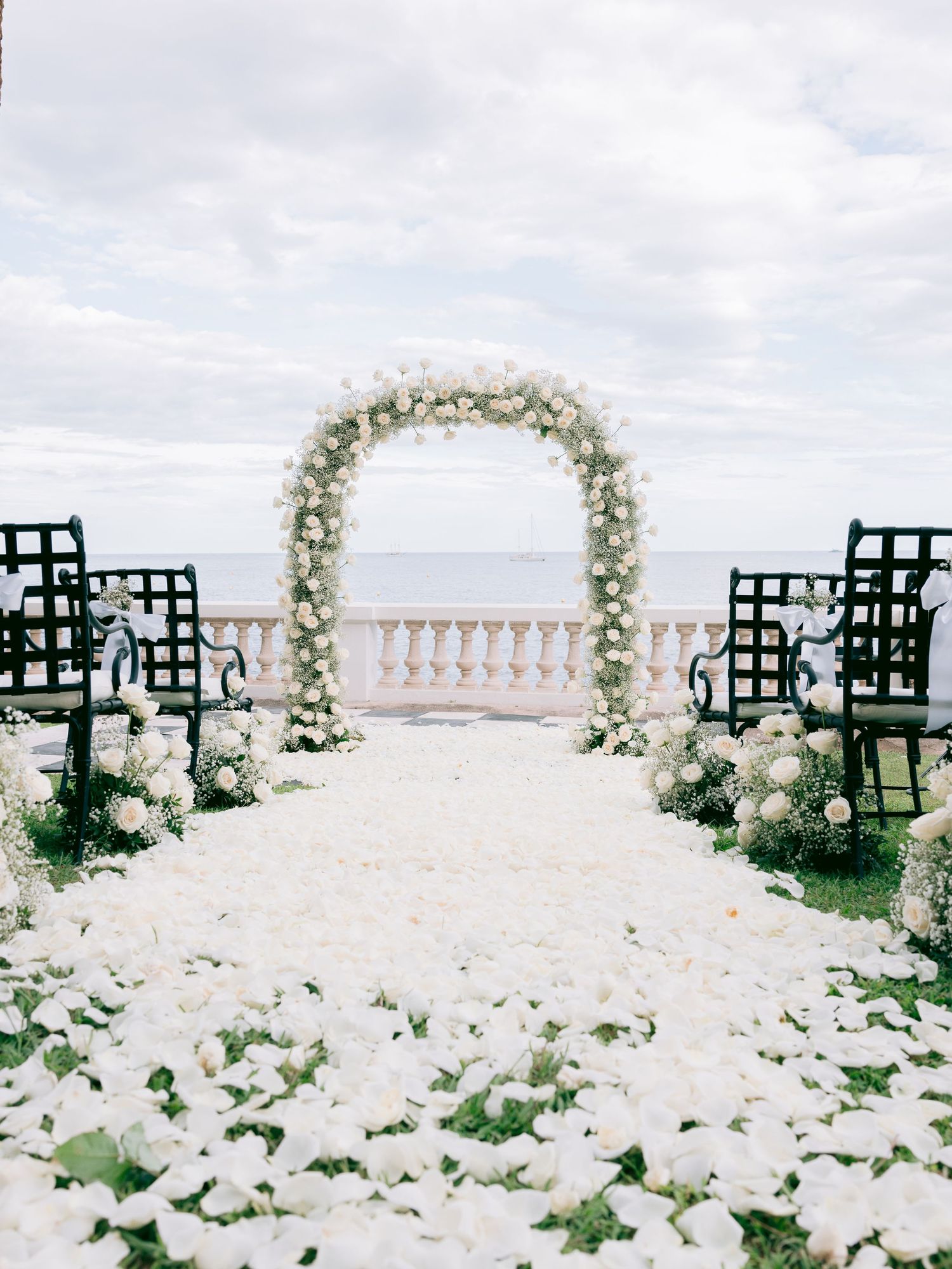 White flower petals line the aisle between rows of black chairs under floral arches by the sea.