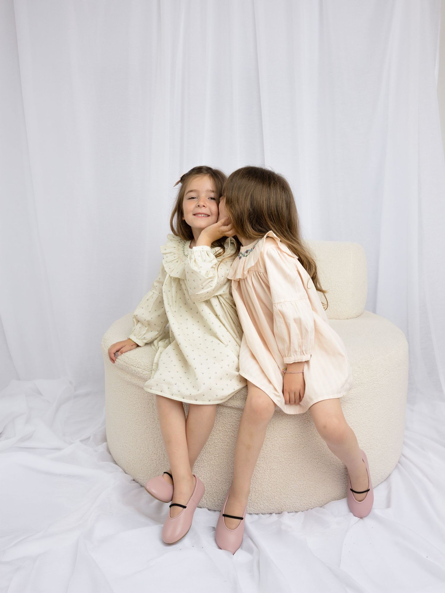 Two young girls in pastel dresses sit together on a cream chair, sharing a sweet moment against a white backdrop.