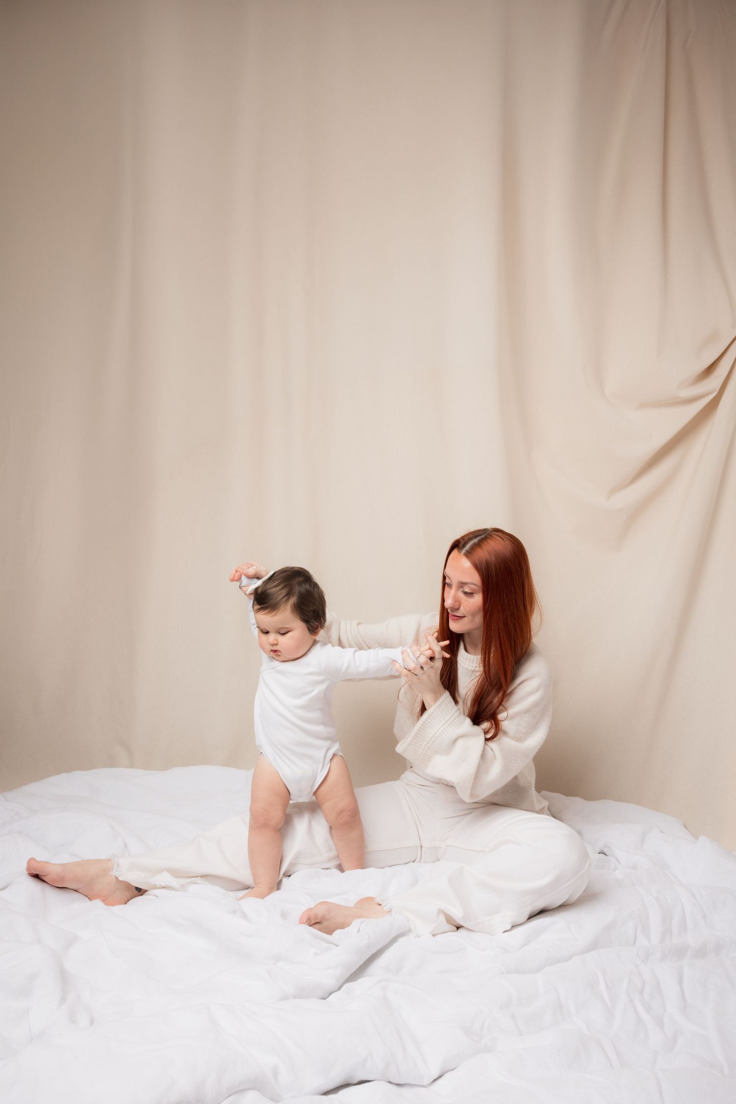Red-haired mother holding toddler's hand on white bed, both wearing white outfits against beige backdrop.