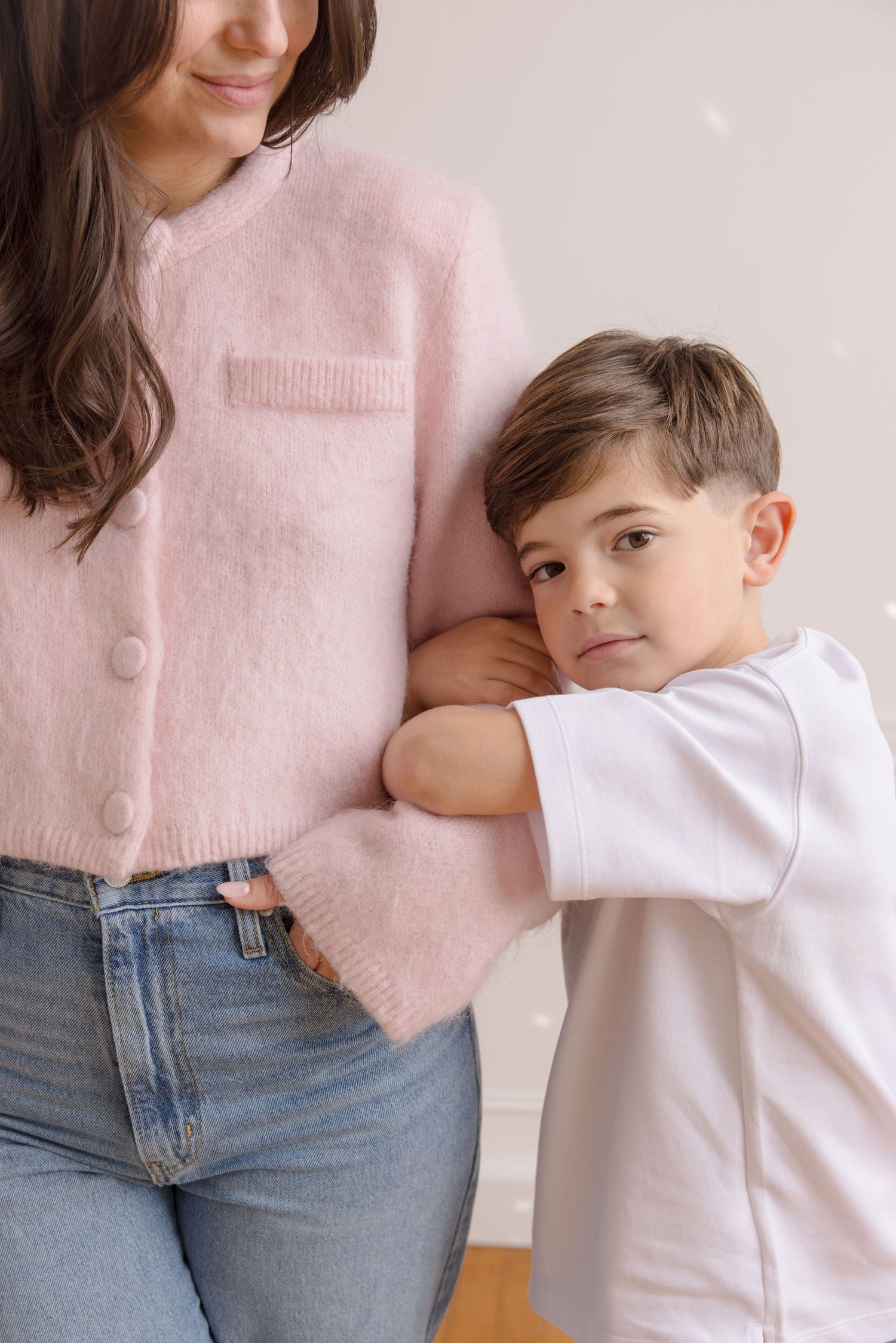 A young boy in a white t-shirt leans against a woman in a pink sweater and jeans in a bright, neutral setting.