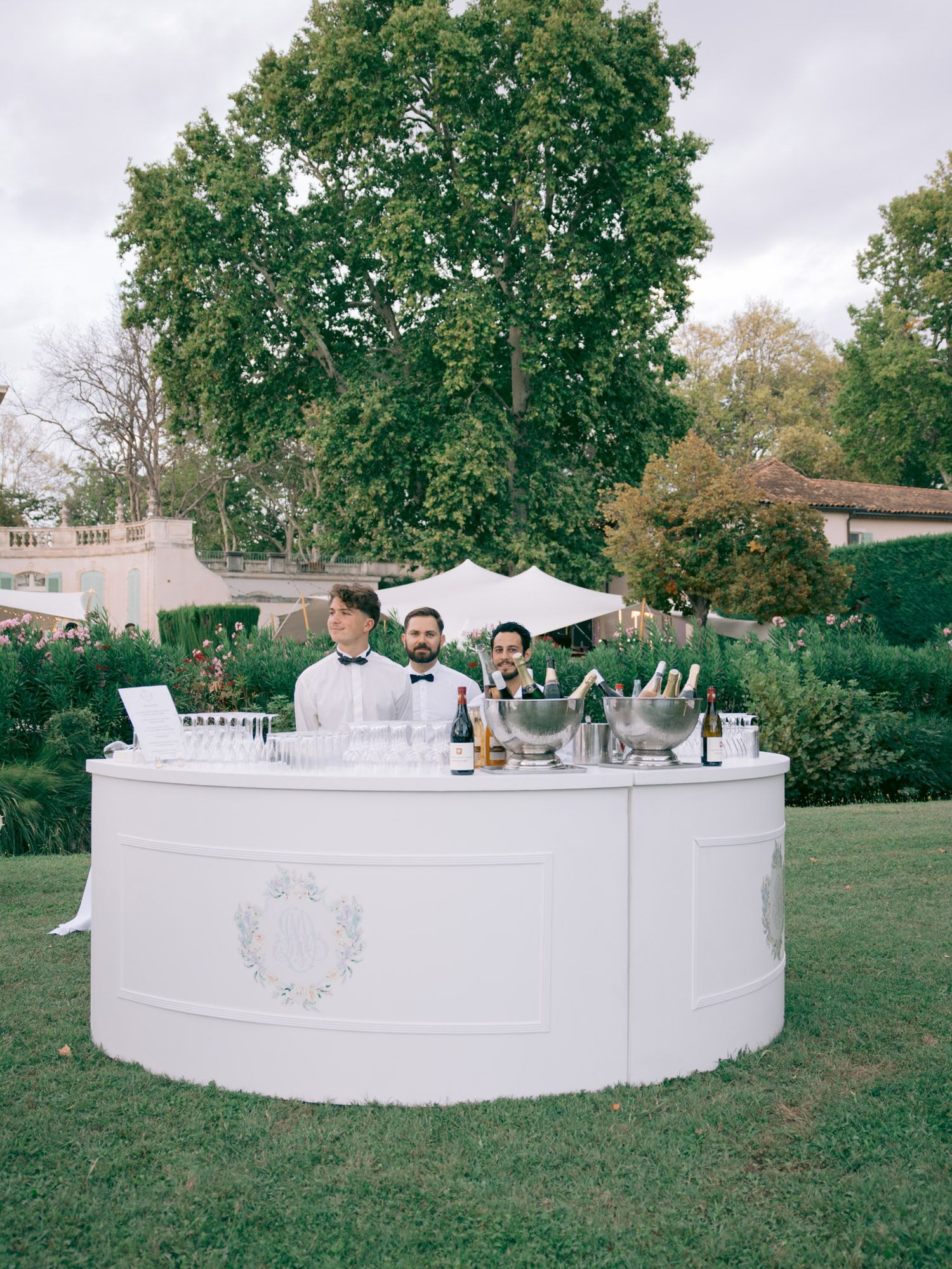 A white curved bar setup at an outdoor wedding reception with servers in formal attire.
