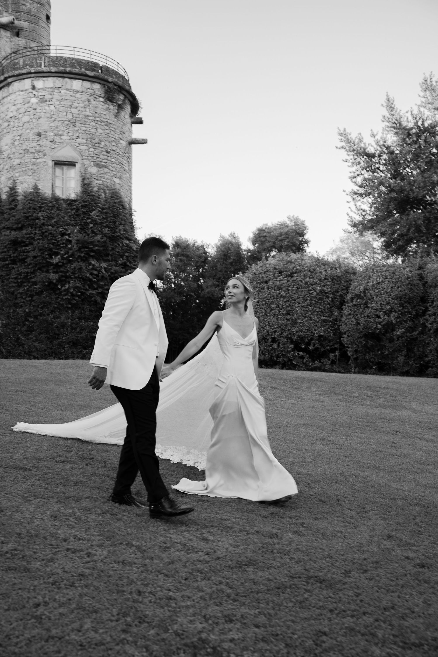 A couple dances together on a grassy lawn near a stone castle wall in black and white.