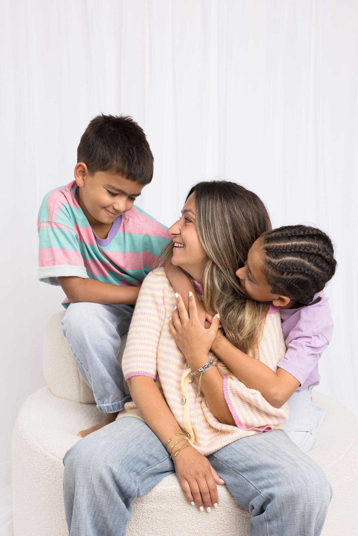 A smiling mother sits with her two children hugging her affectionately against a white curtain backdrop.