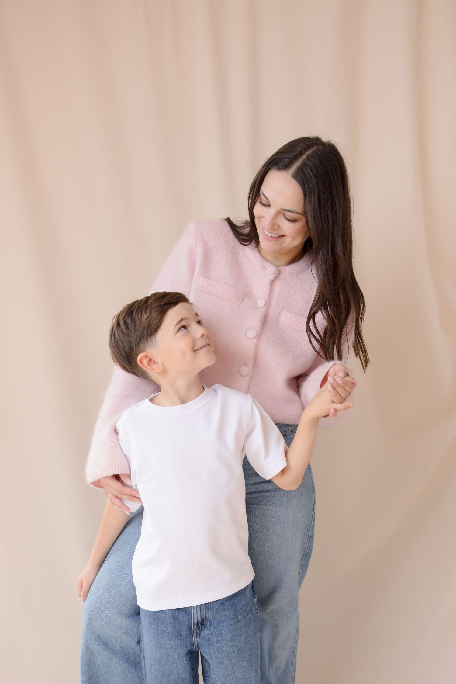 A smiling woman in a pink jacket and jeans stands with a young boy in a white t-shirt against a beige background.