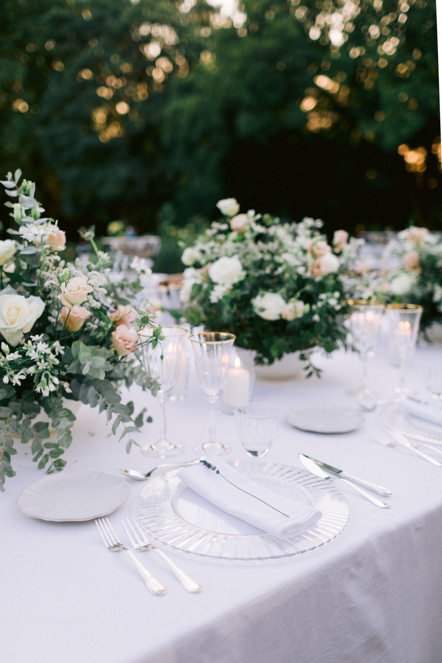 Elegant table settings with white linens, floral arrangements and silverware for an outdoor wedding reception.