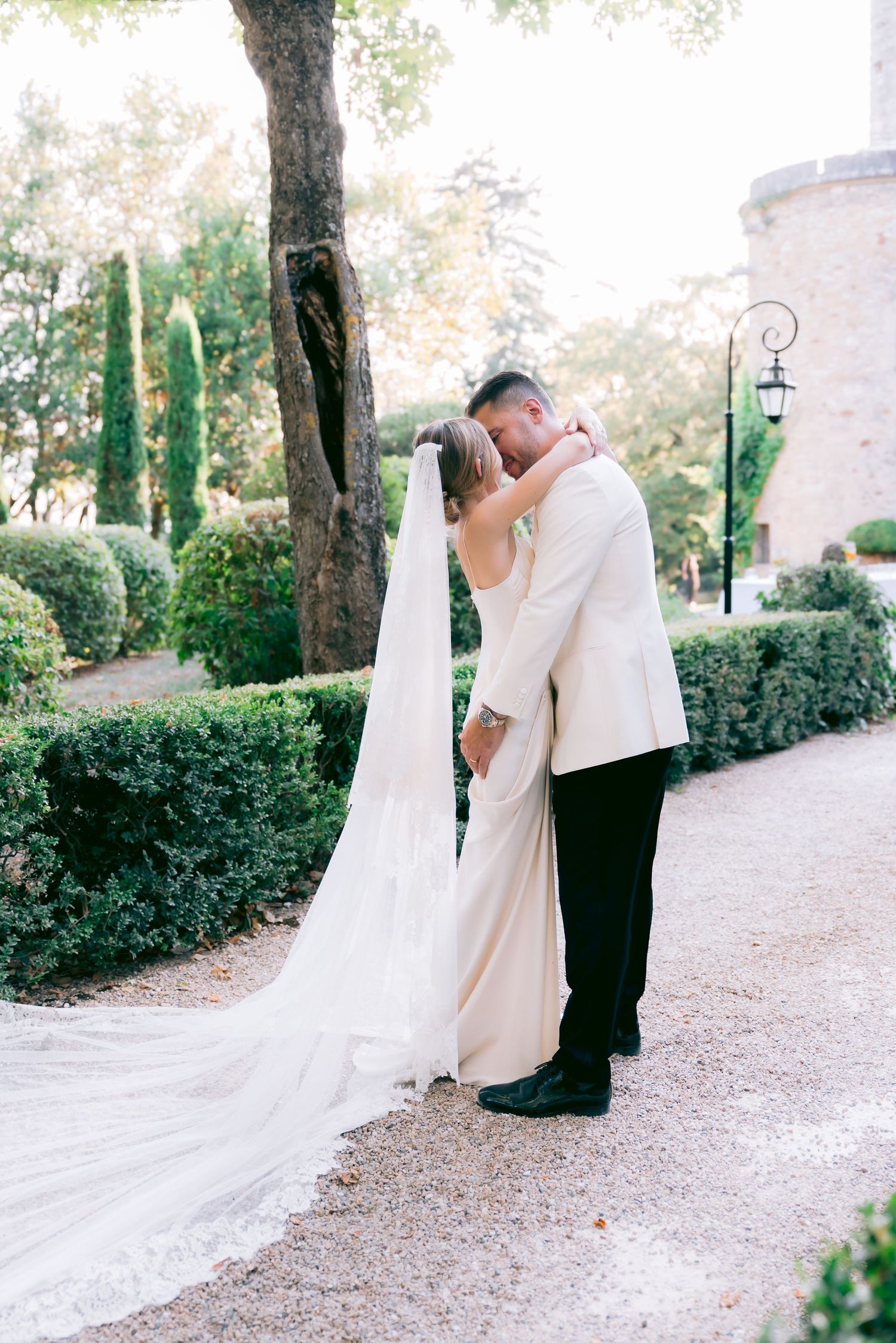 A romantic wedding couple shares an intimate moment in a garden path lined with cypress trees and greenery.