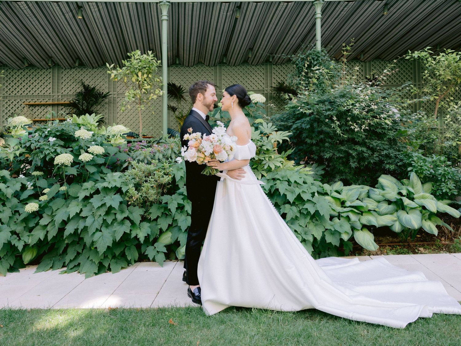 Wedding dress train trails along lush garden pathway surrounded by greenery.