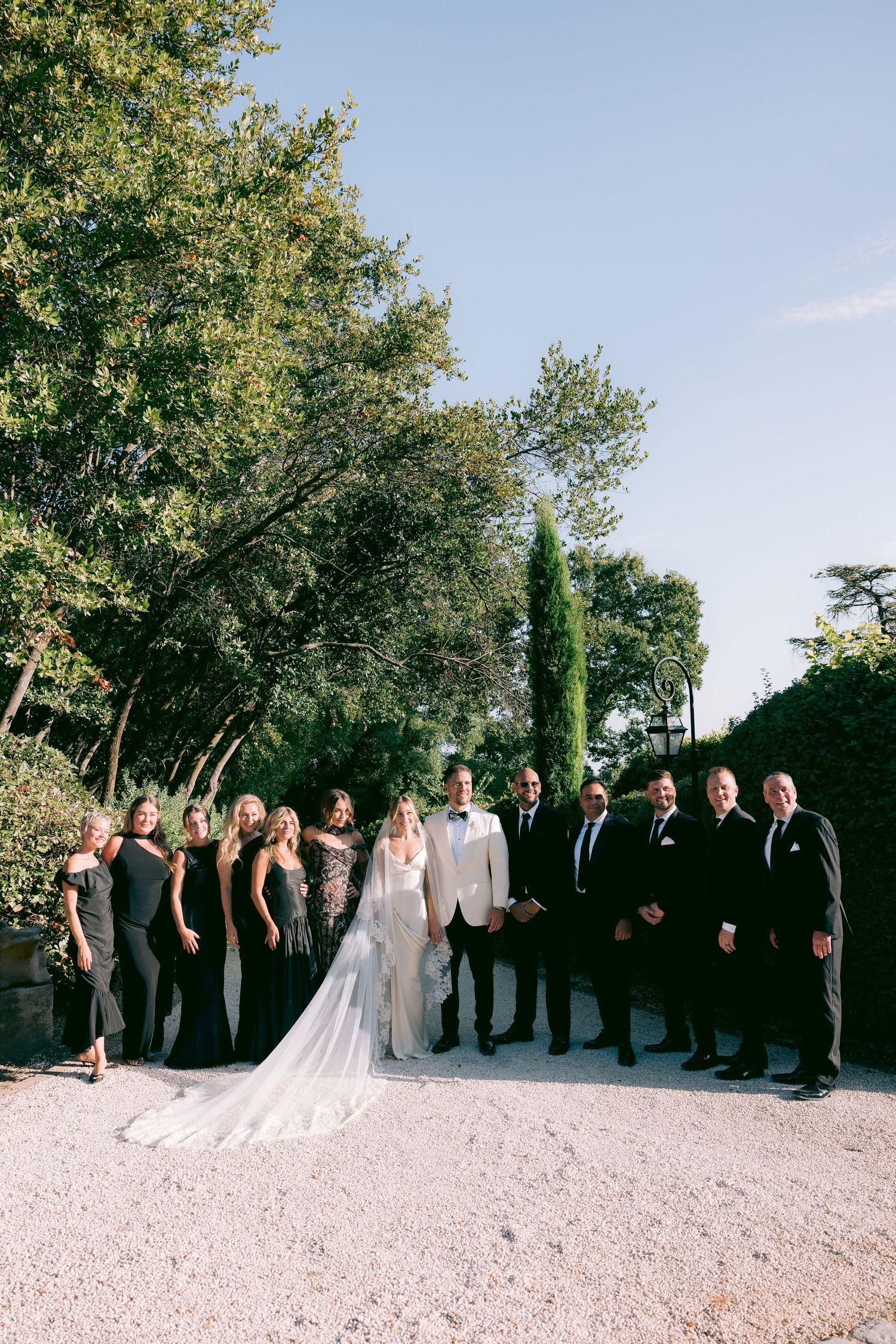 Wedding party poses together outdoors under green trees on a sunny day.