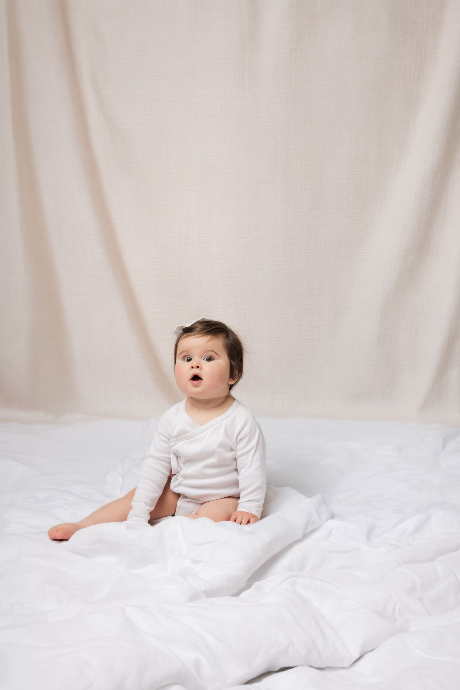 Cute baby in white onesie sitting on white bedding against a soft beige fabric backdrop.