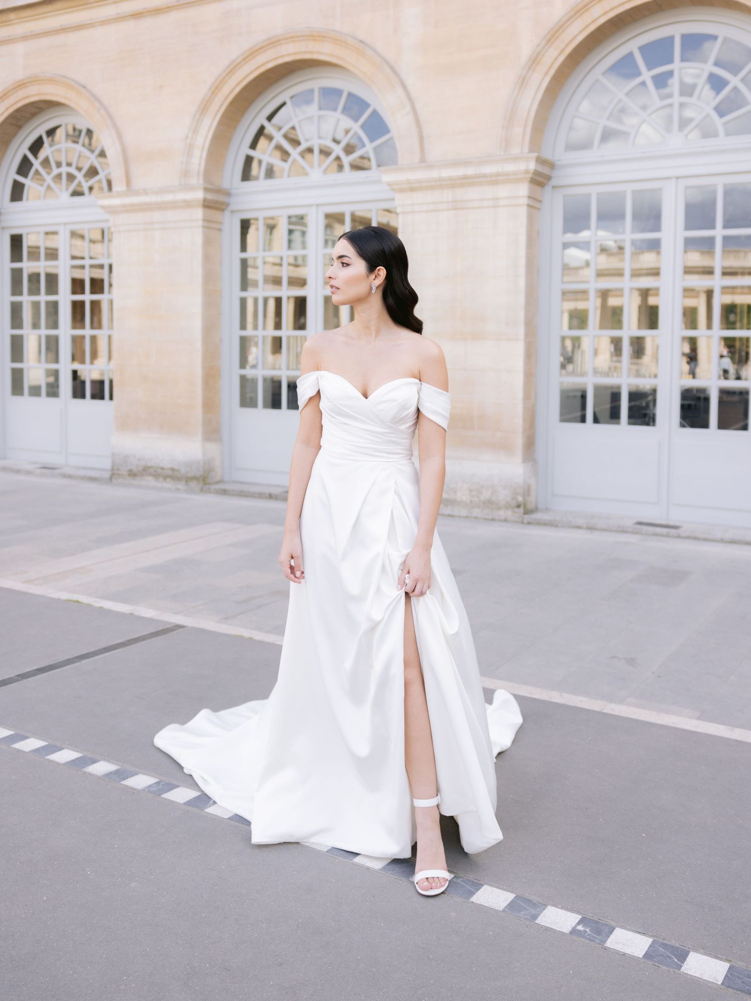 Elegant bride in a flowing white wedding dress walks gracefully across a historic building exterior with arched windows.