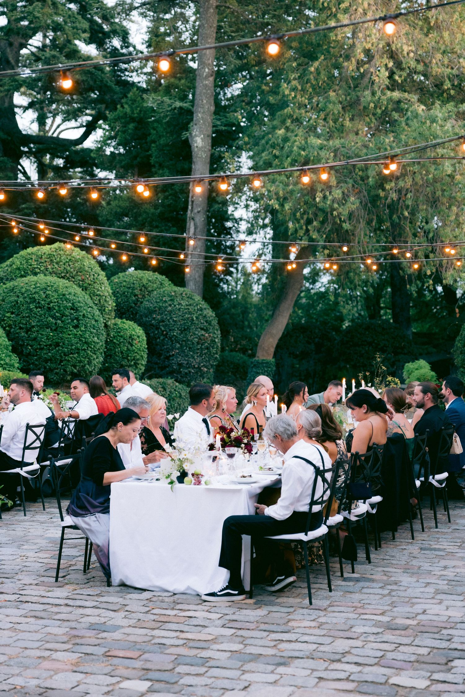 Outdoor wedding reception with string lights and guests dining at long tables on a stone patio.