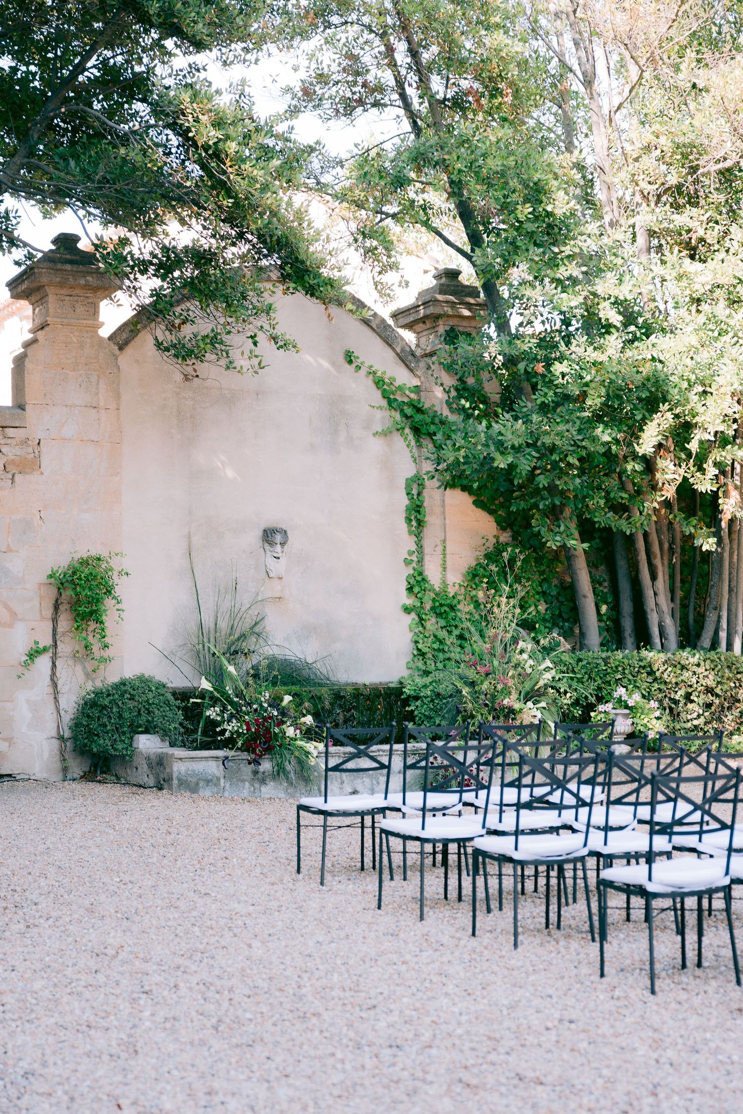 Outdoor wedding venue with stone walls and metal chairs arranged on gravel ground beneath overhanging trees.