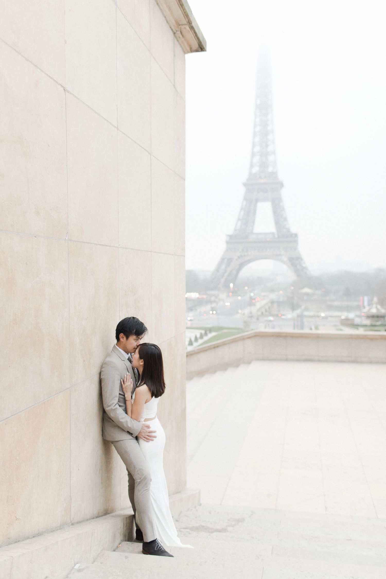 Romantic couple embracing against stone wall with Eiffel Tower in misty background.