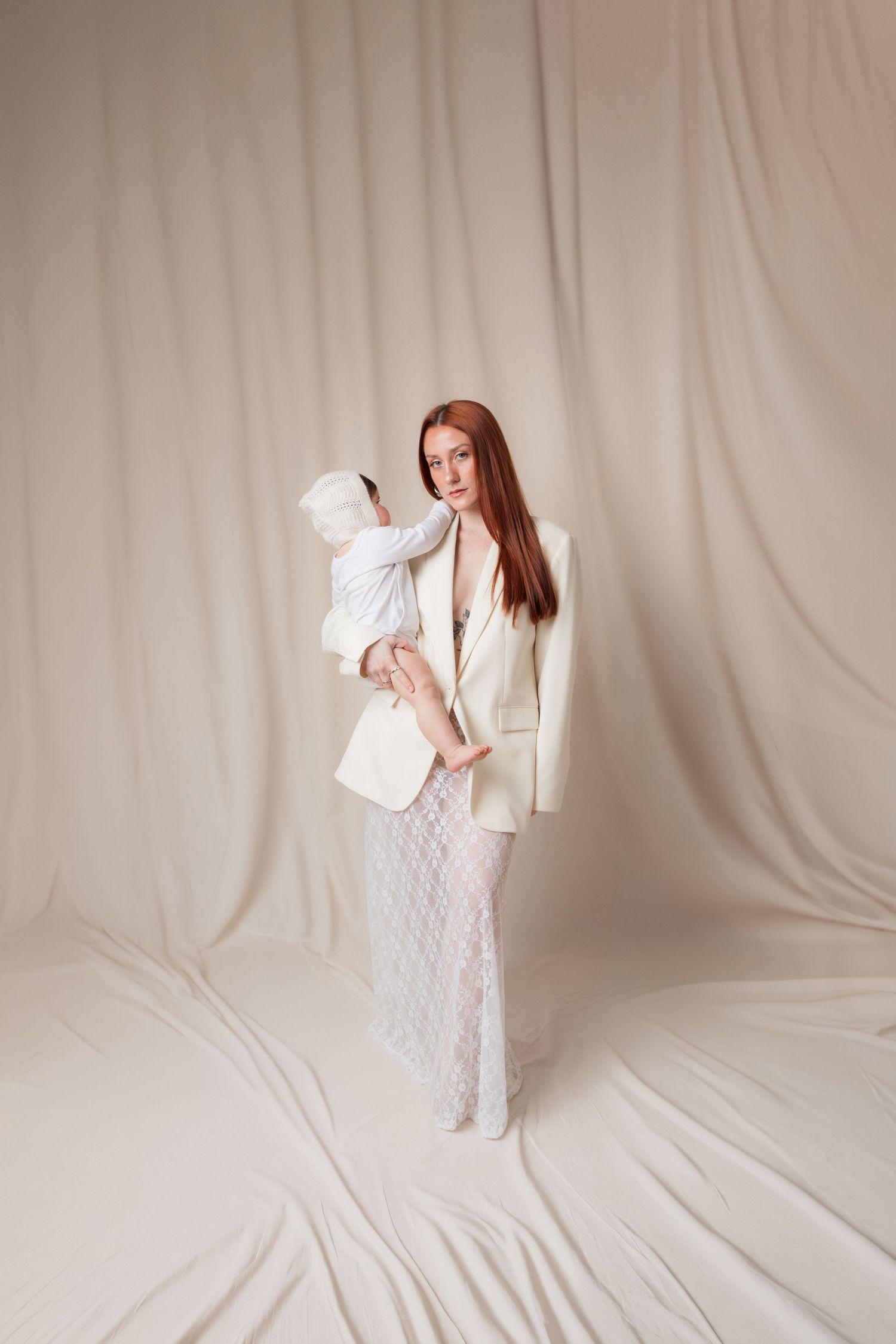 A redheaded woman in a white blazer and lace skirt holds a baby against a soft draped cream fabric backdrop.