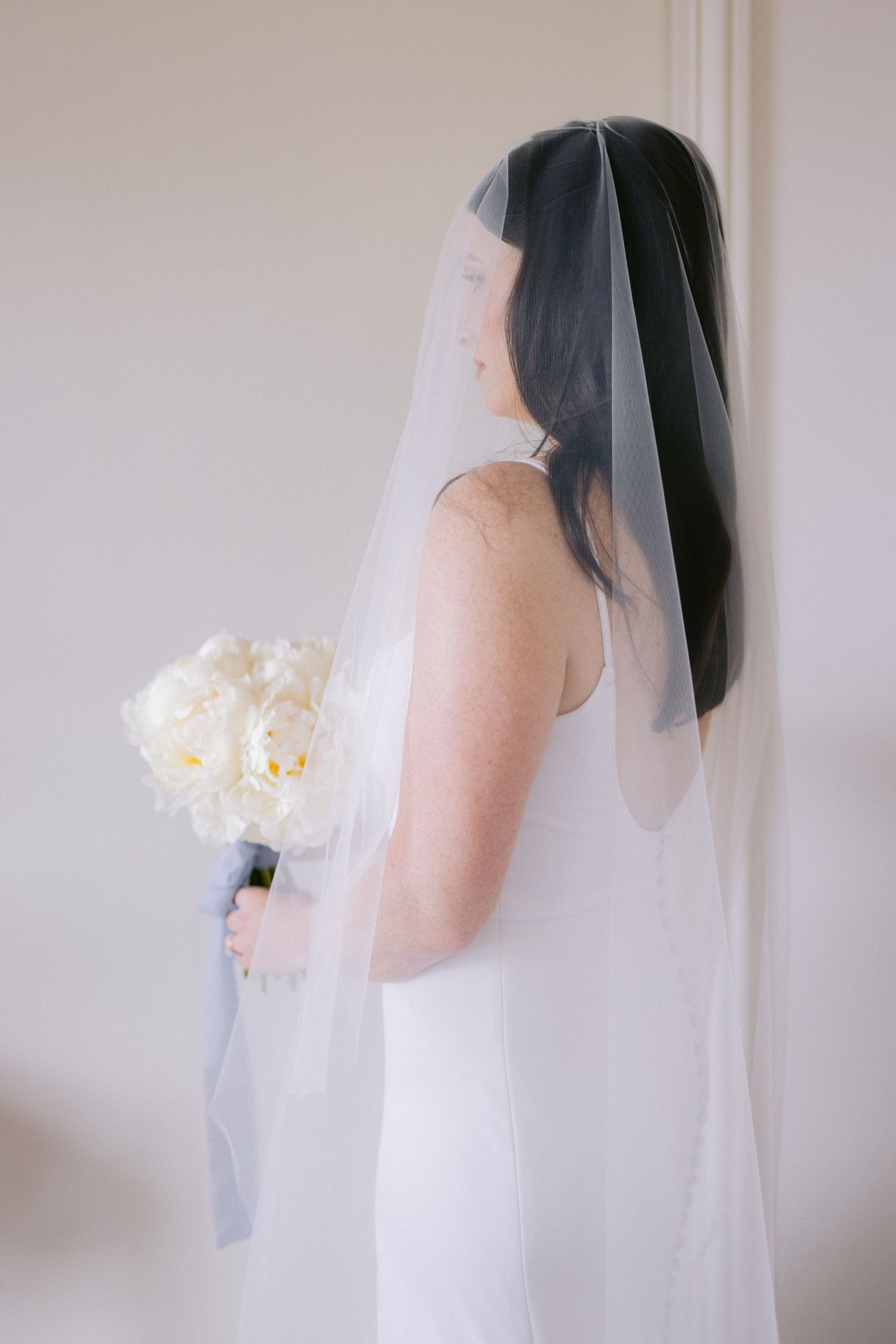 Side profile of bride in simple white dress with veil and peony bouquet.