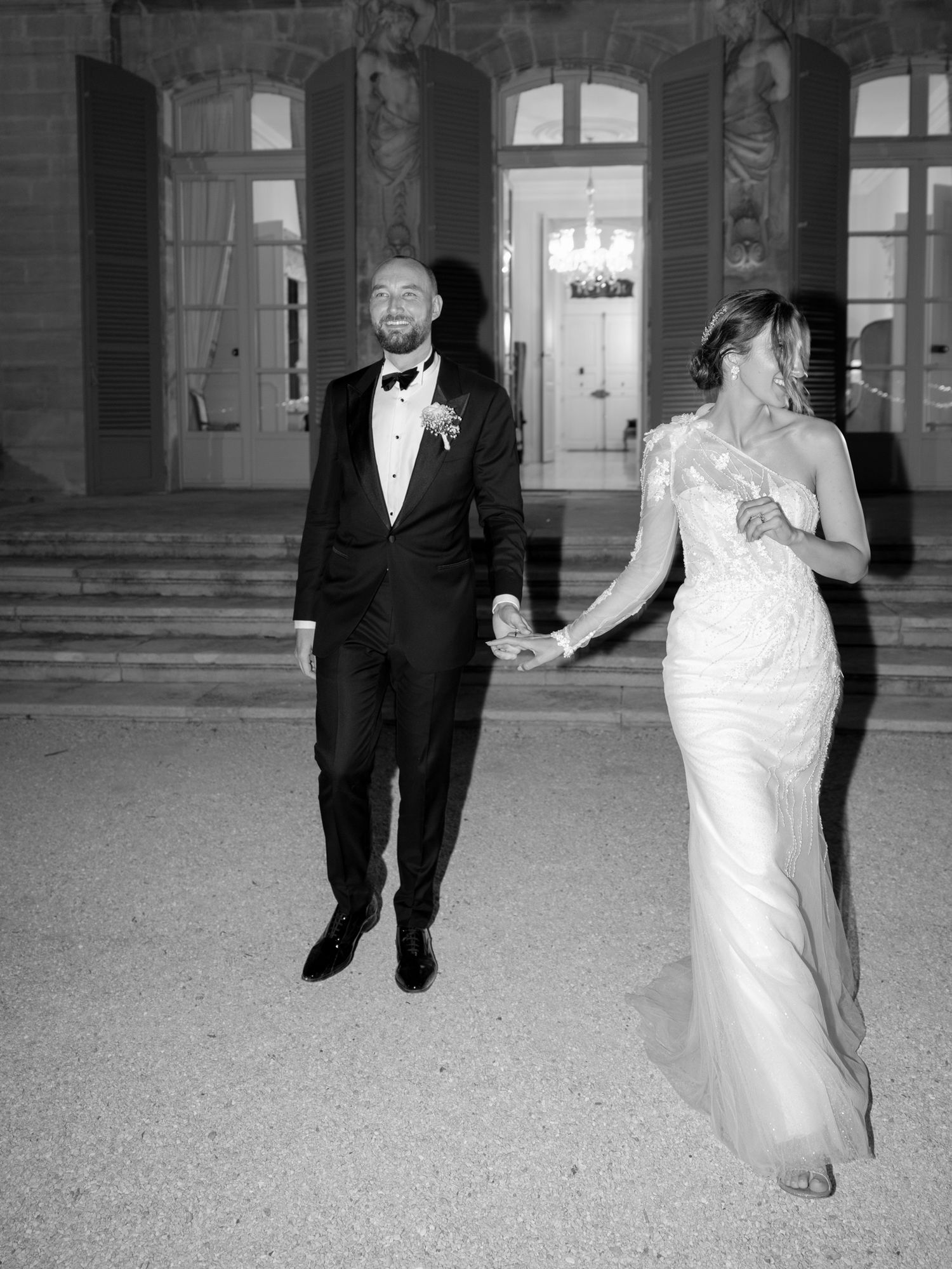 A black and white photo captures a couple in formal wedding attire walking outside a grand building.