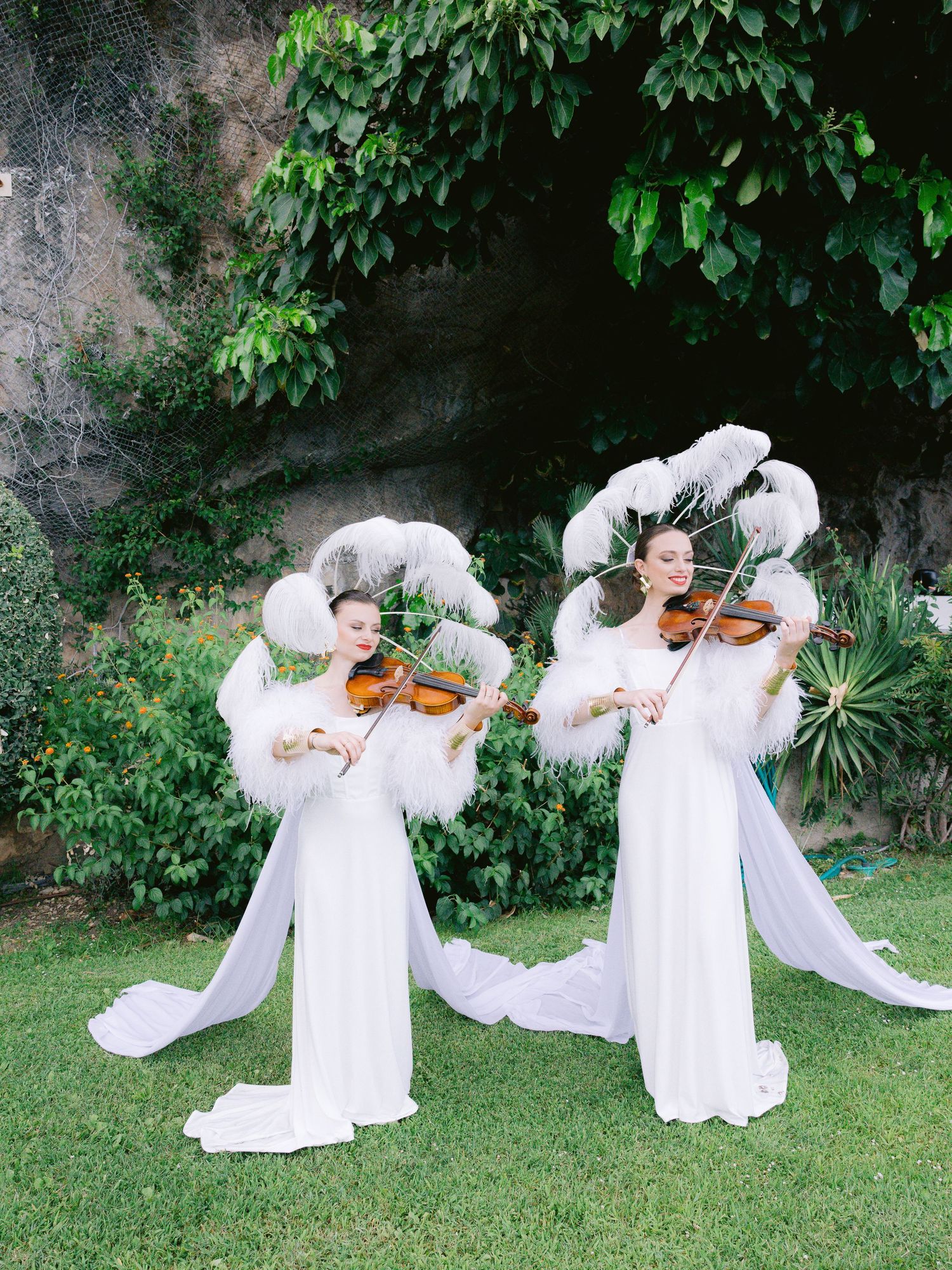 Musicians in white dresses performing with violins in a garden setting.