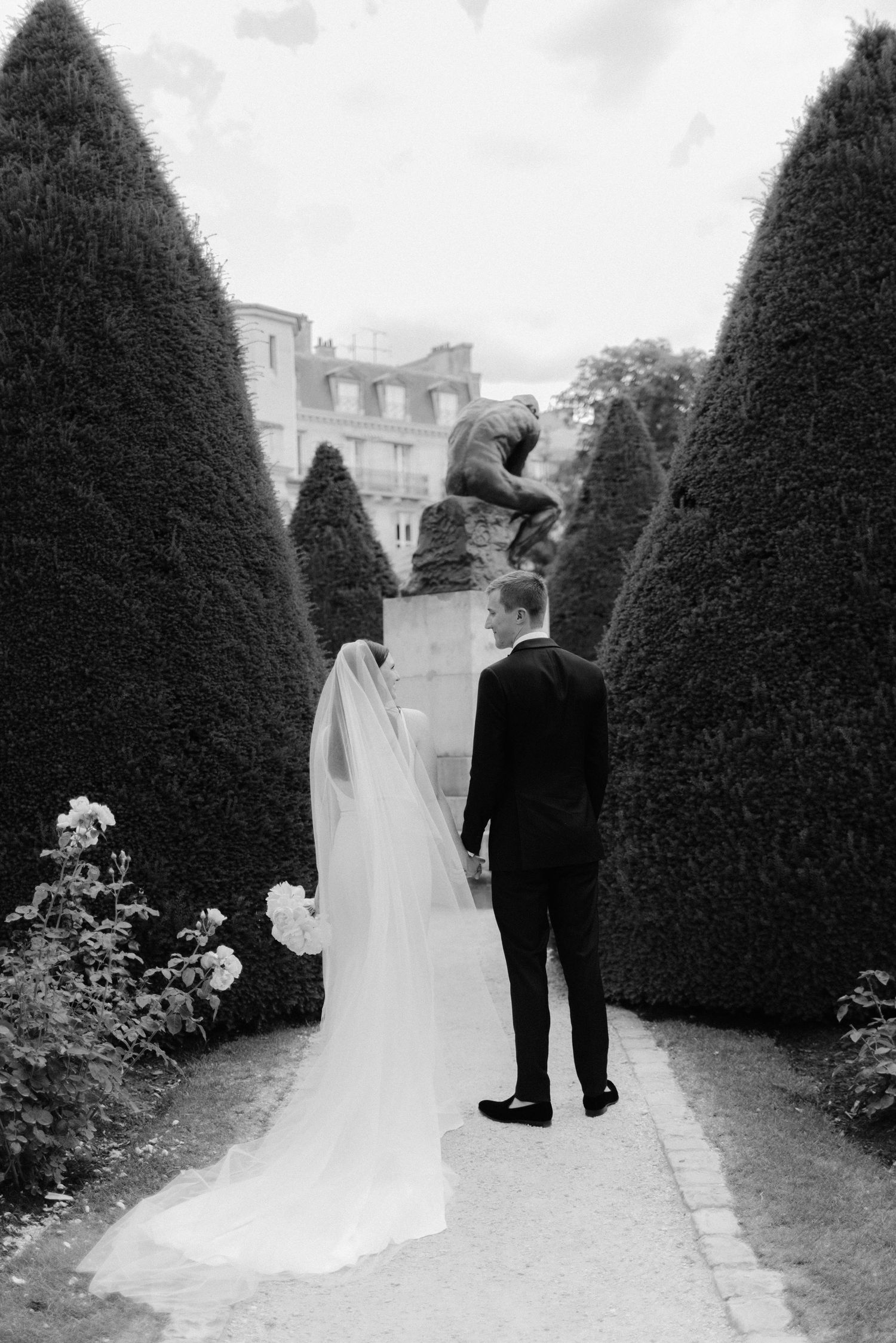 A couple walks through a garden path lined with perfectly manicured topiary trees.