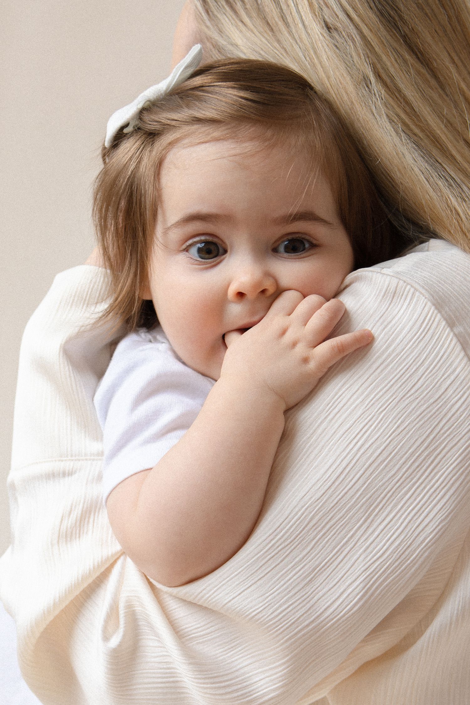 Adorable baby girl with brown eyes resting on mother's shoulder, hand near mouth, wearing white onesie.