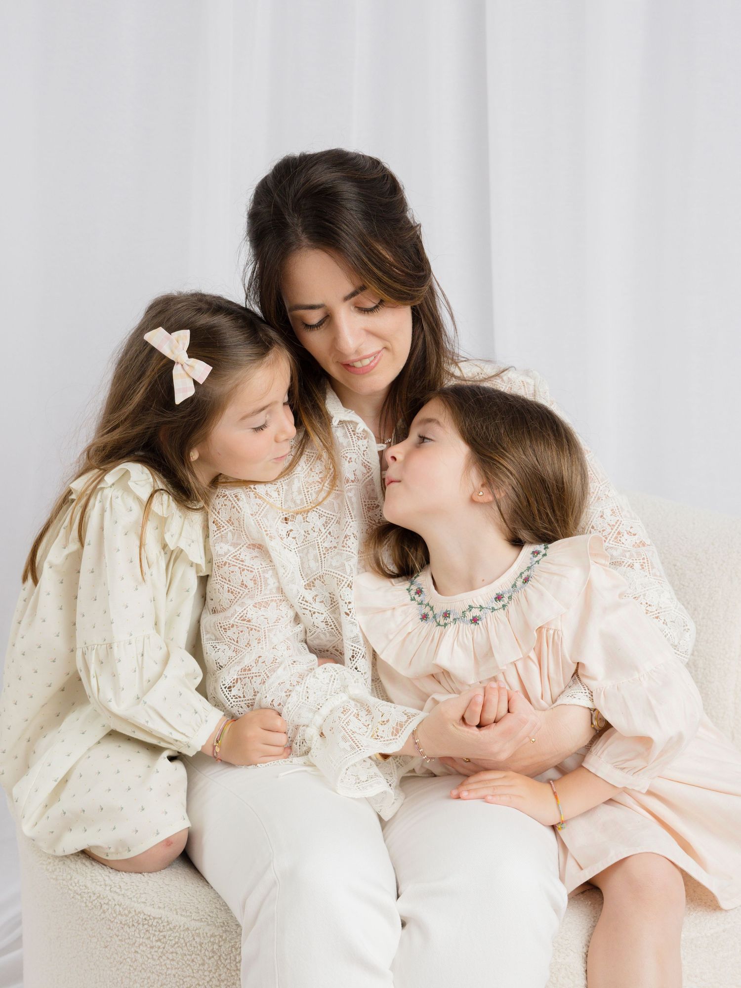 A mother in white lace sits with her two daughters in pastel dresses, sharing a tender embrace on a cream sofa.