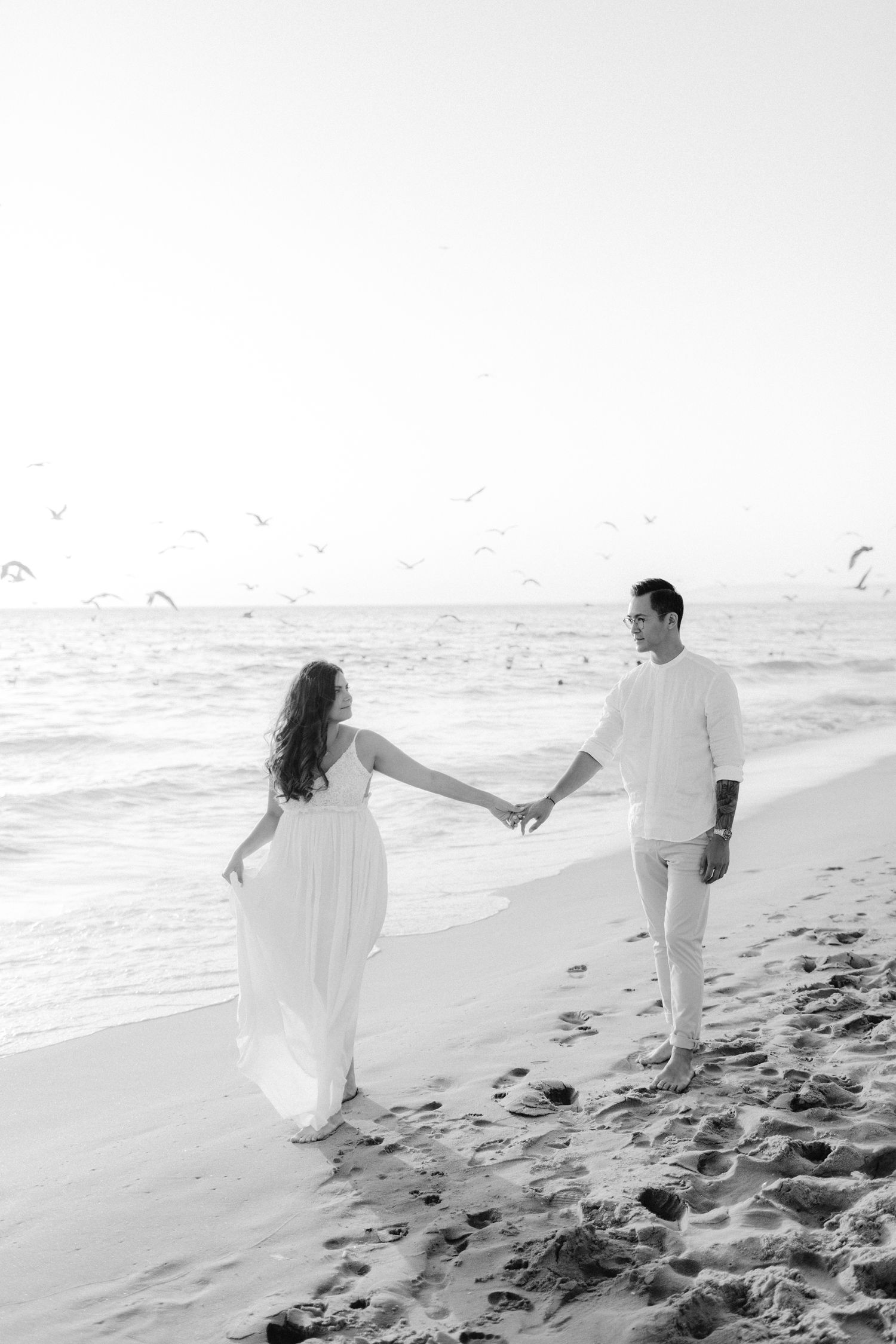 Couple holding hands while walking along beach shore in black and white.