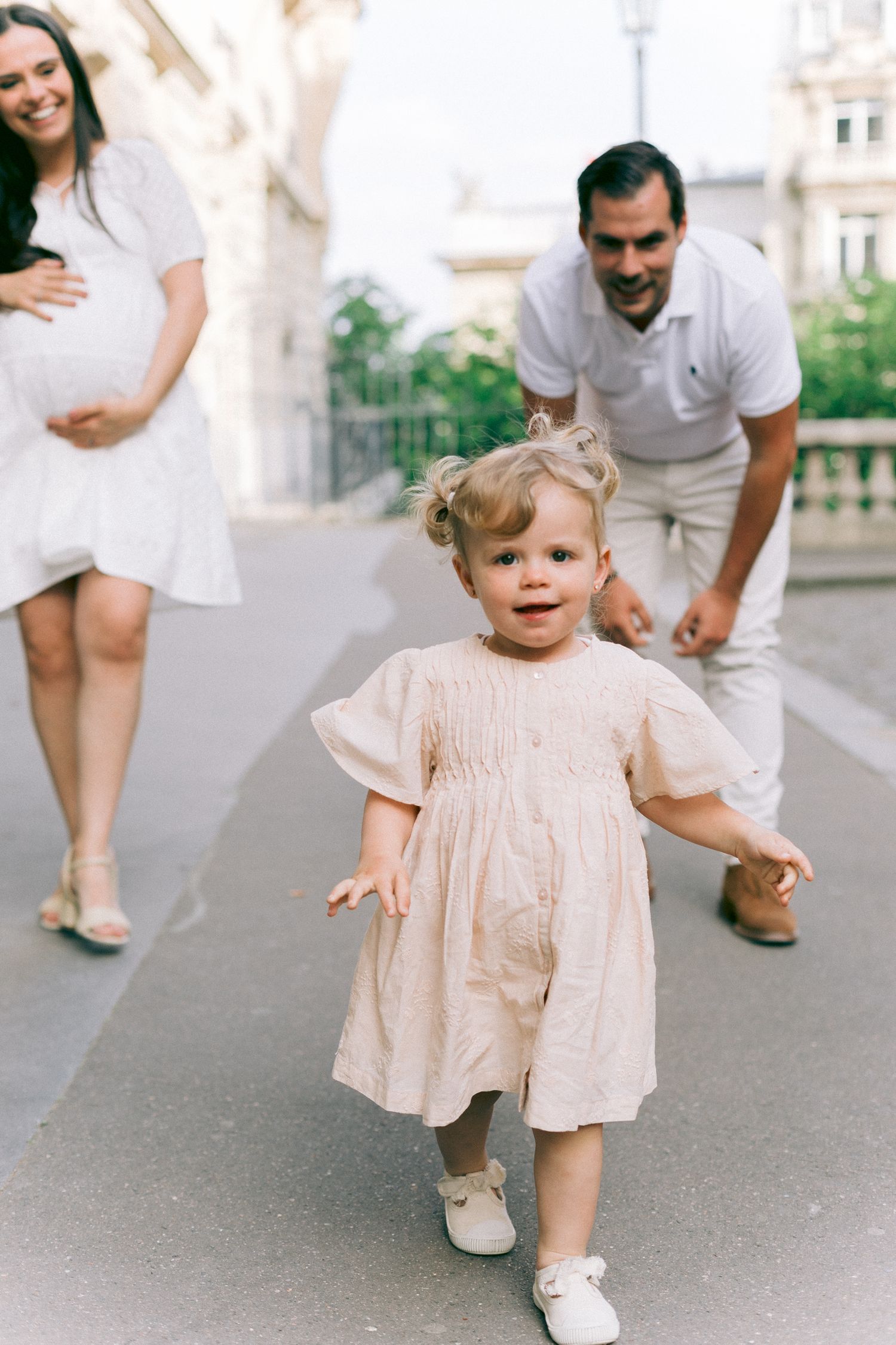 A toddler in a pink dress takes joyful steps on a city street while adults watch from behind.