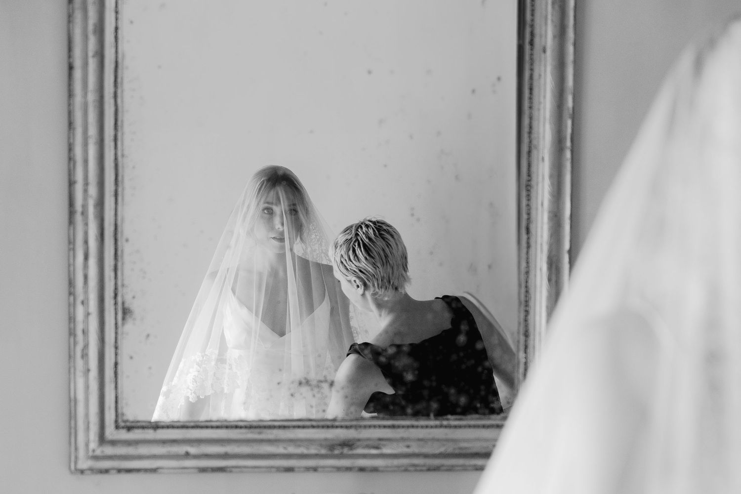 Black and white photo of a wedding dress and veil in a room with ornate mirror.