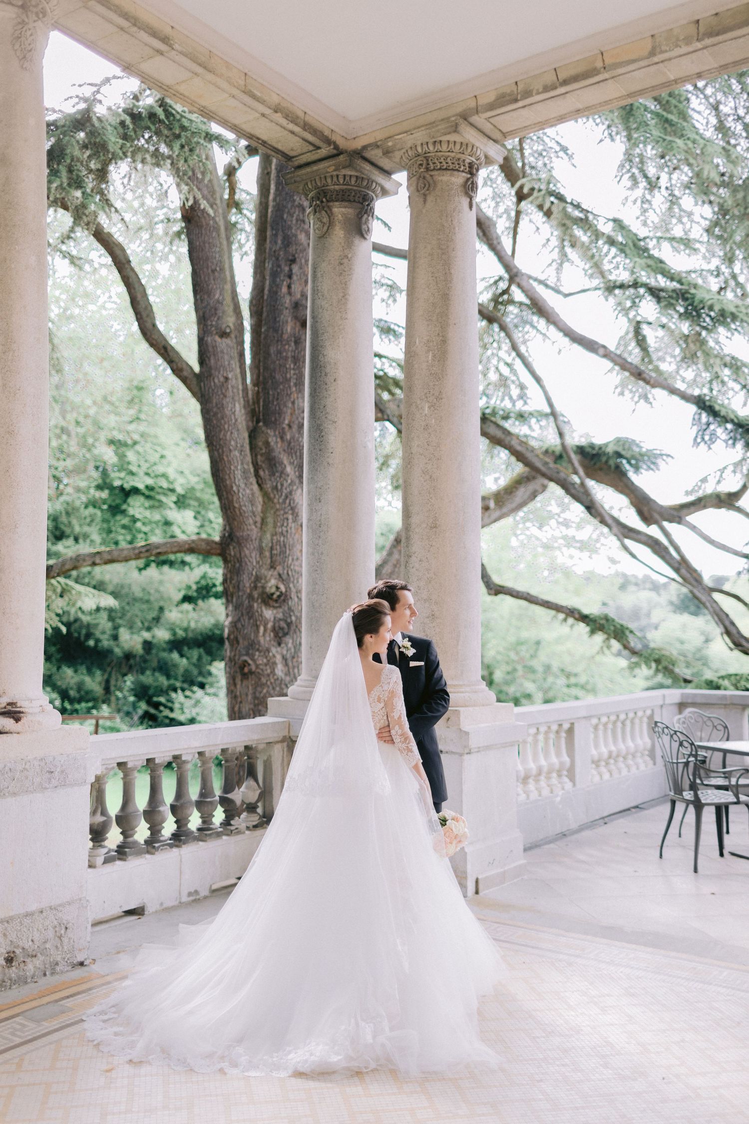 Wedding couple photographed on an elegant columned terrace surrounded by trees.