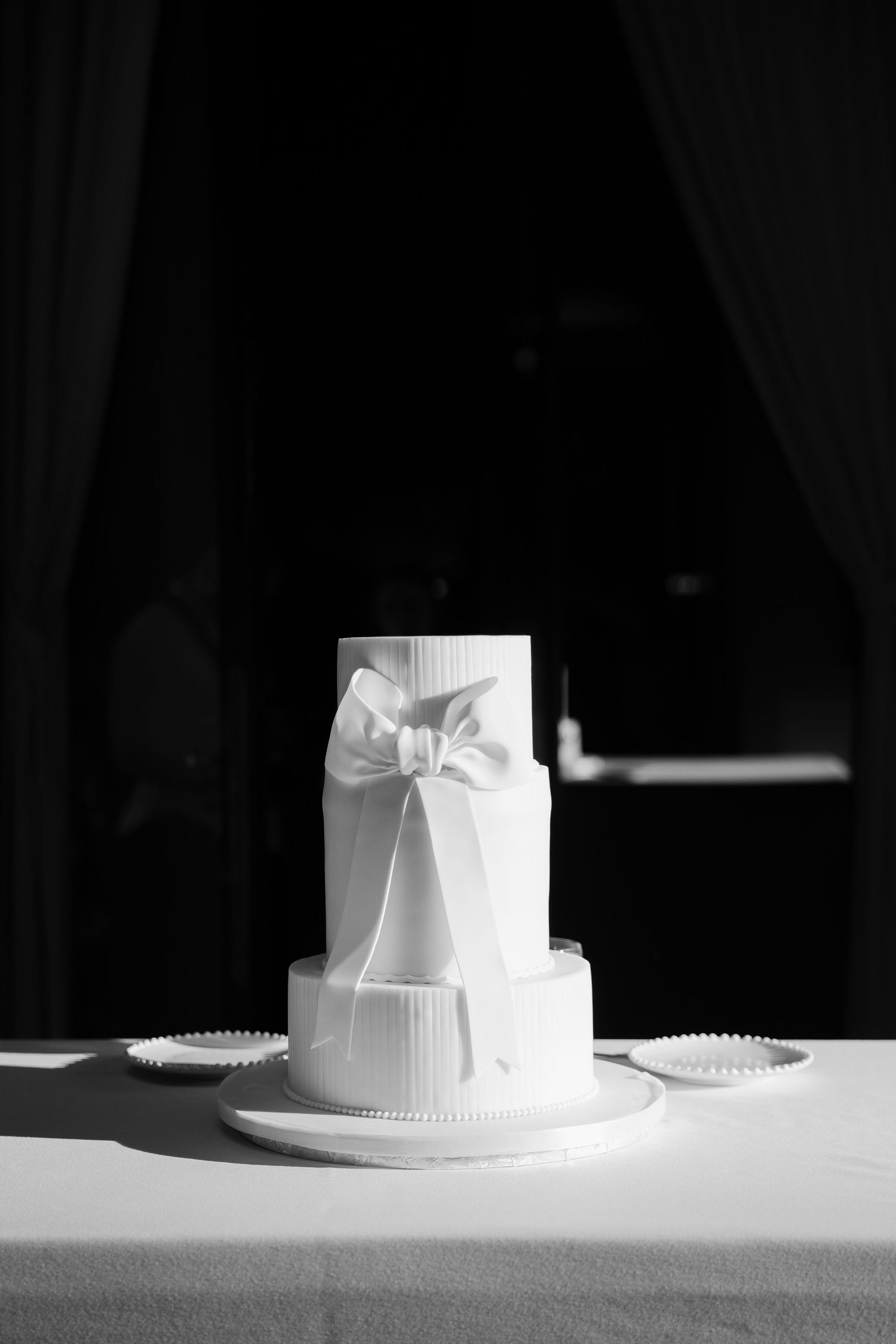 Minimalist white wedding cake with elegant bow detail photographed in dramatic lighting.