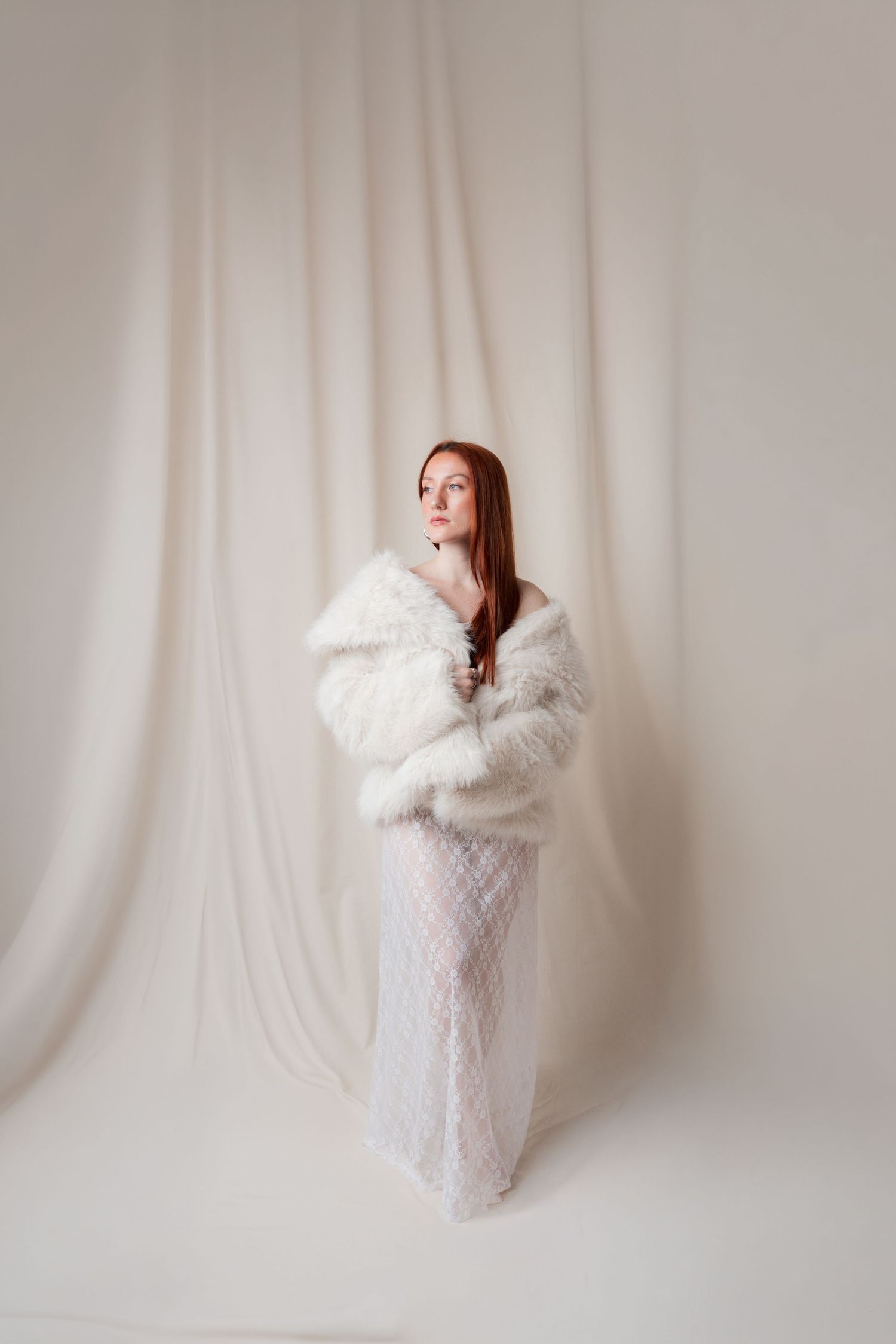 Young woman with red hair wearing a white fur coat and lace dress, posing elegantly against a soft white draped backdrop.