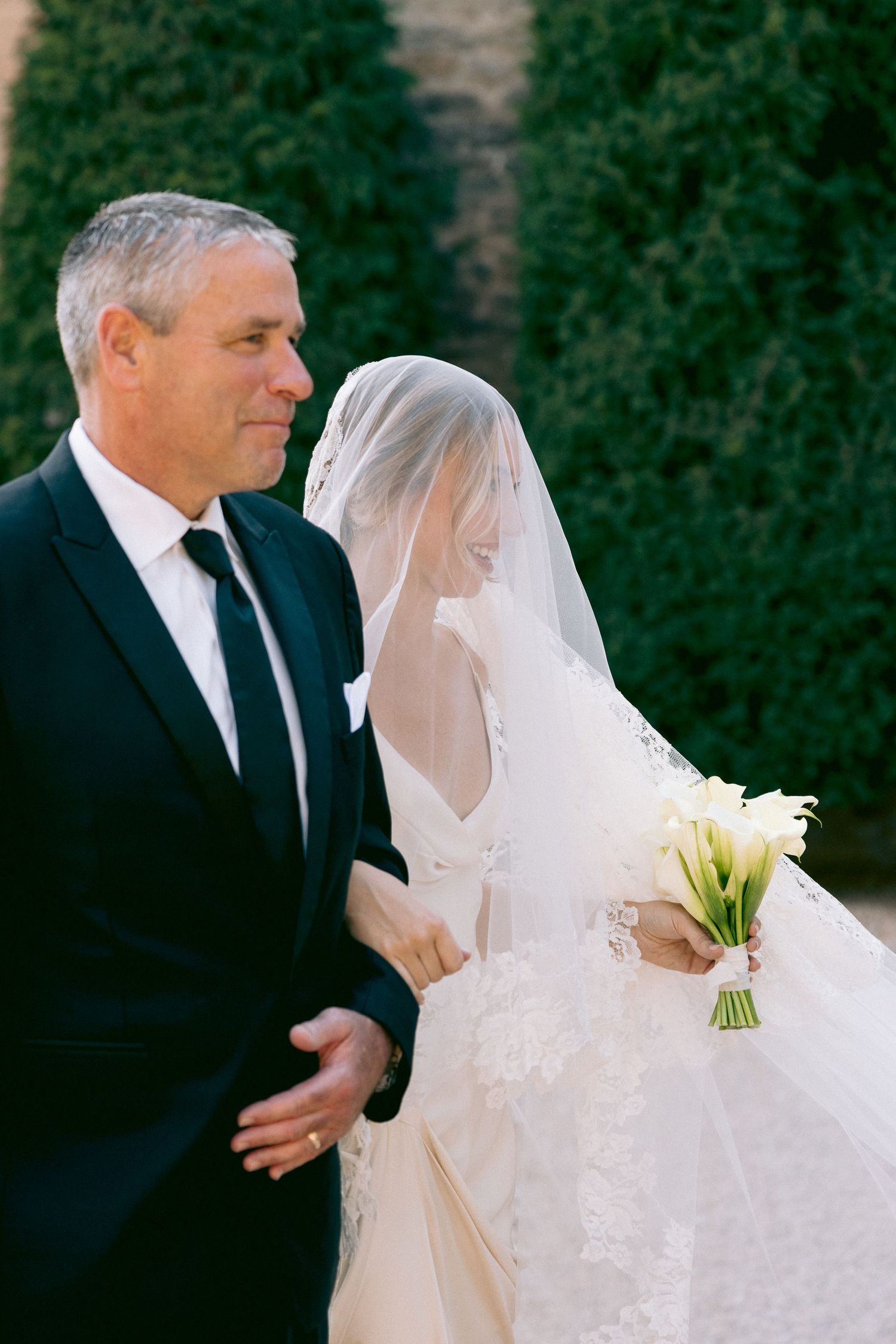Wedding couple walking along stone wall with lush green topiaries and white bridal bouquet.