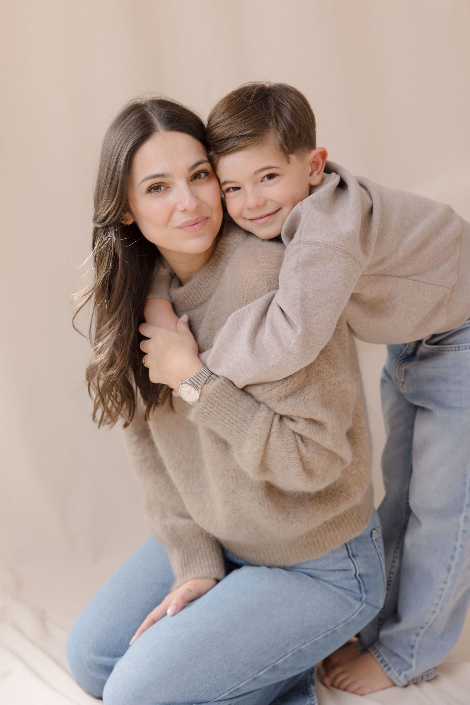 A woman and young boy in matching beige sweaters and jeans share a warm hug during a studio photo session.