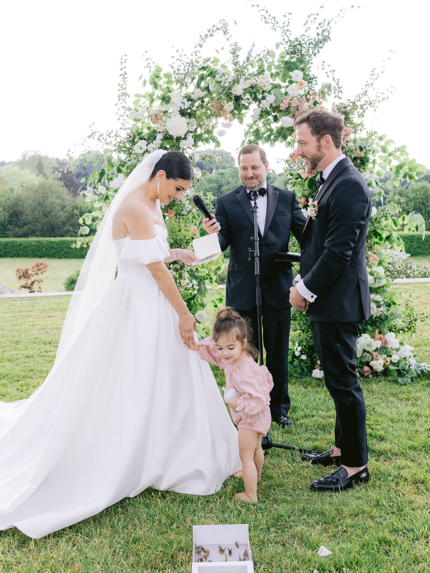 A whimsical outdoor wedding scene with guests gathered around floral decorations.