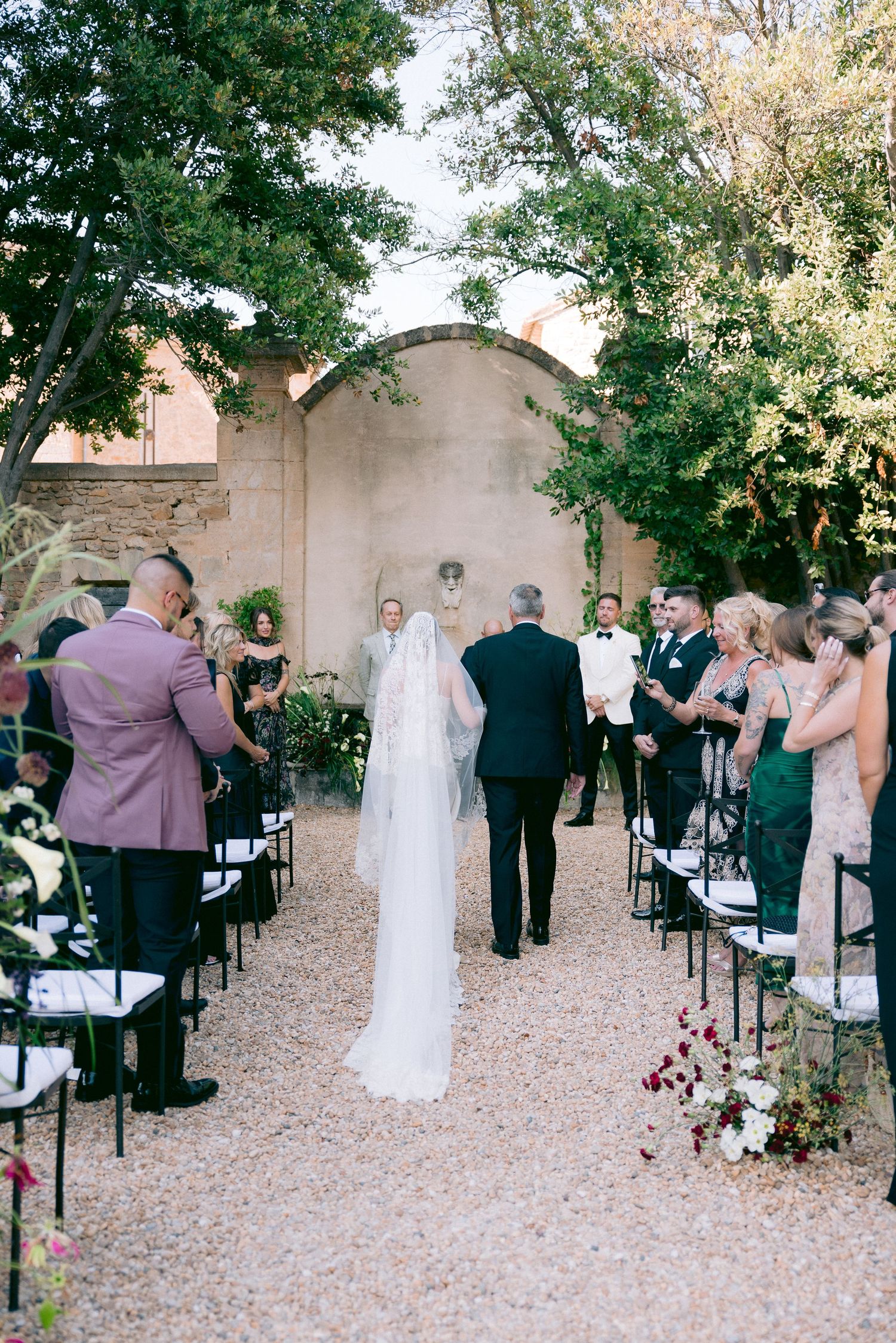 Wedding guests gathered in outdoor courtyard with chairs arranged for ceremony under trees.