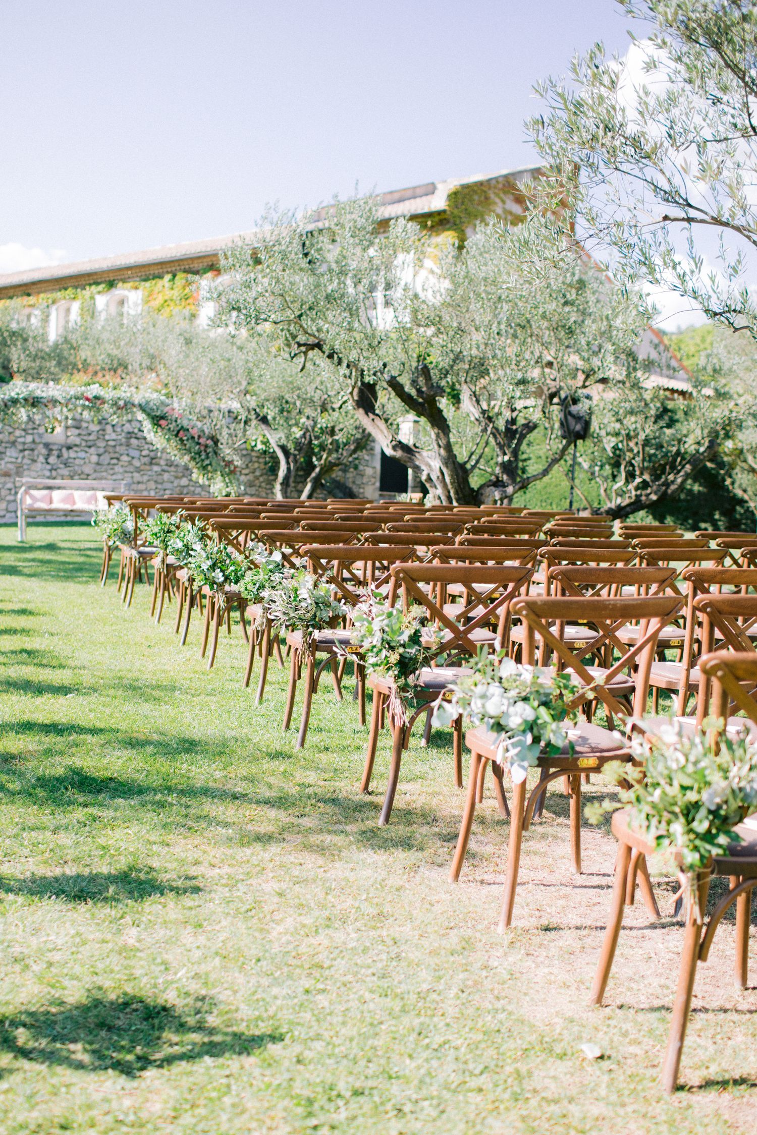 Wooden ceremony chairs decorated with floral arrangements line up in rows on a grassy outdoor venue.