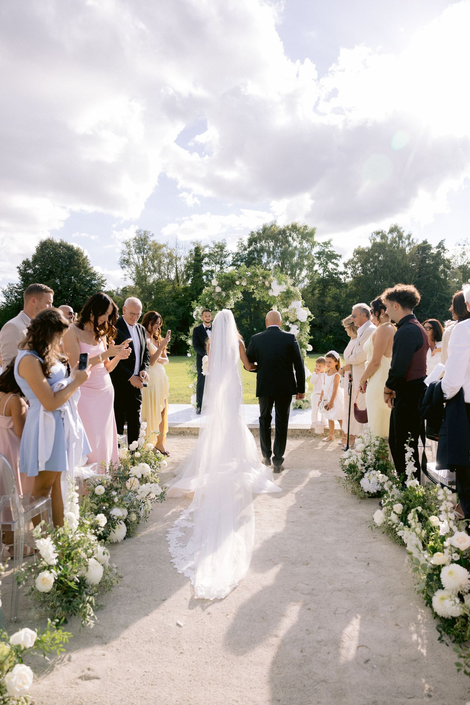 Château de Sept Saulx Wedding ceremony guests gather outdoors along a decorated path with white floral arrangements.